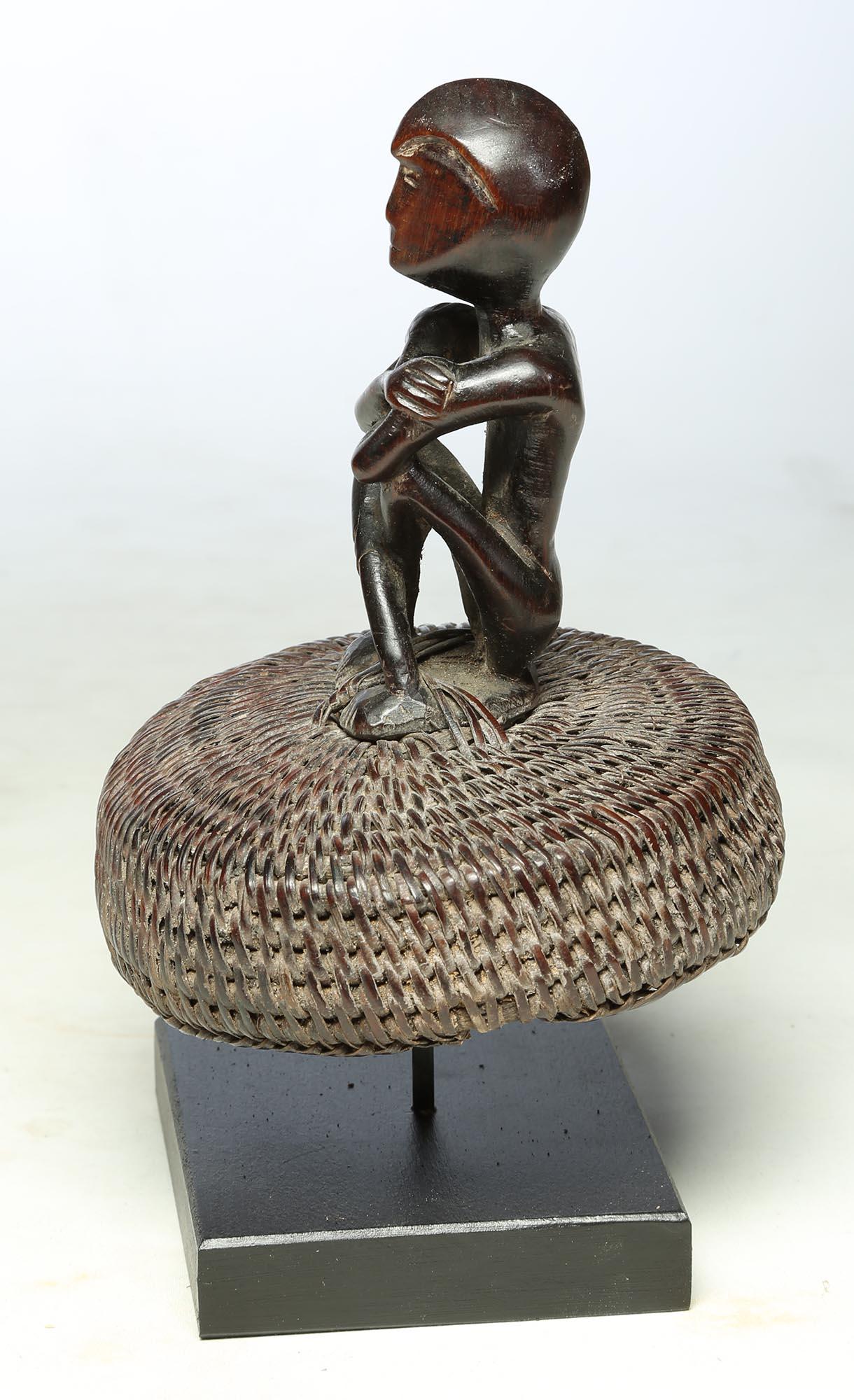 Carved wood seated Philippine miniature Bulul figure sitting and attached to the top of an old hat or basket lid with crossed arms. Worn and polished surface. Measure: figure alone 4 1/2 inches high, figure & basket 6 1/2 inches, on custom wood and