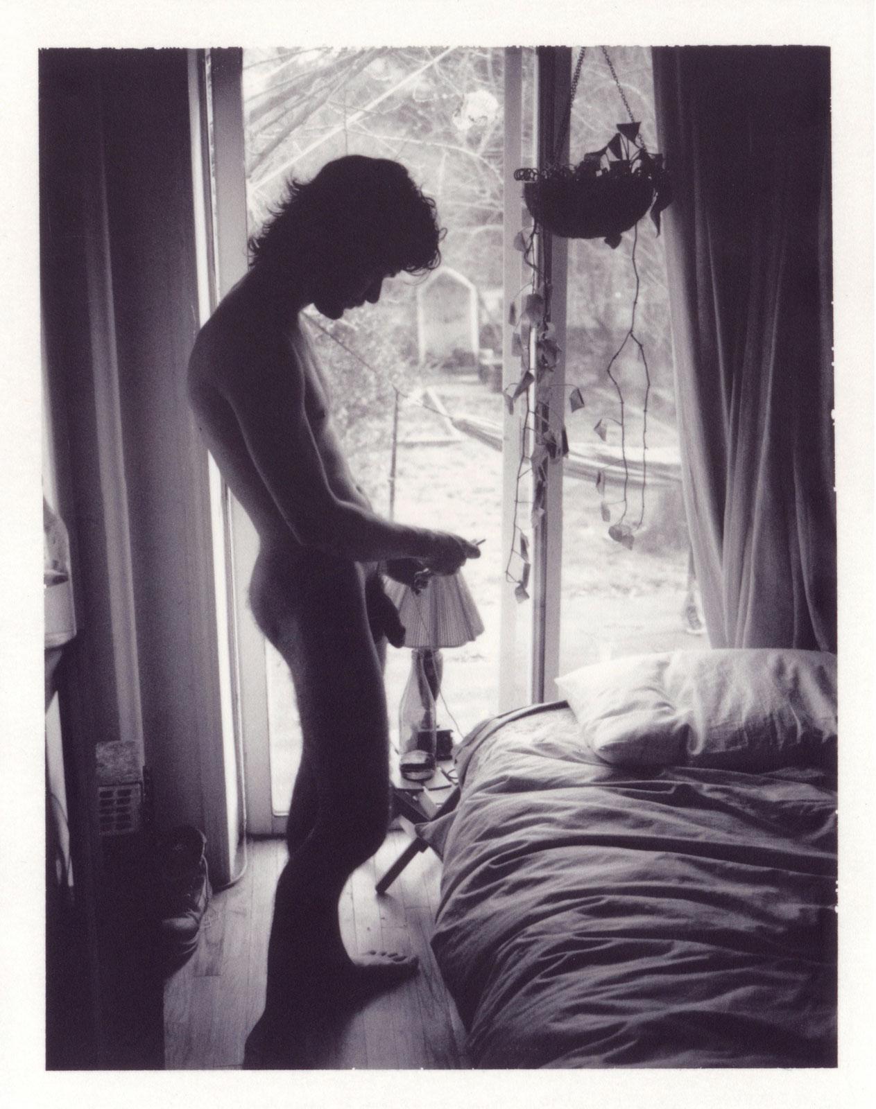 Tanner's Room in Greenpoint (Young, hung nude man checks iPhone by unmade bed) - Photograph by Sebastian Perinotti