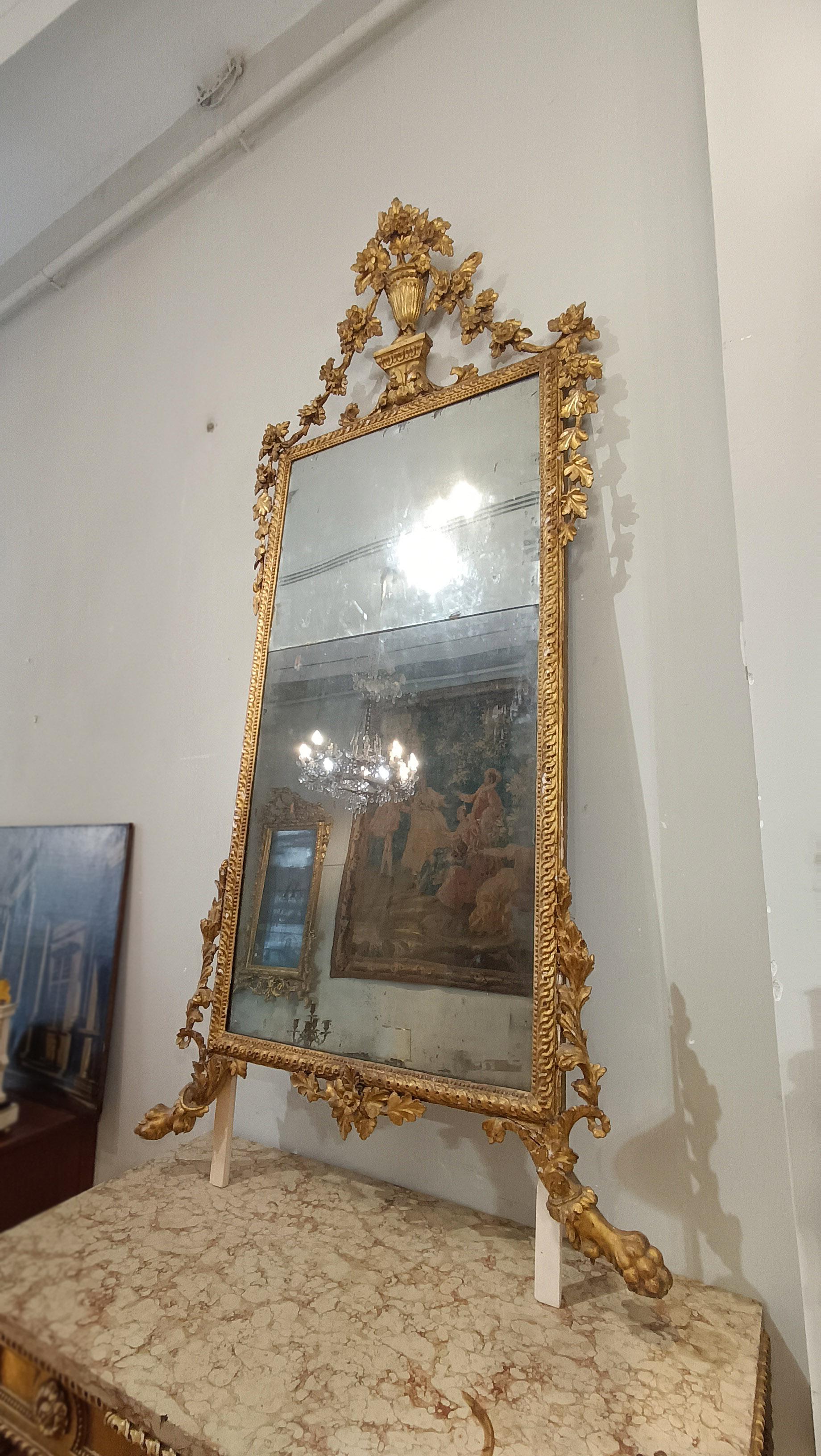 Elegant and imposing mirror with a pine wood frame, finely carved and gilded with pure gold leaf. The frame is adorned with sophisticated guilloches and beads, while the lower part ends with feral legs and plant decorations. The cymatium also stands