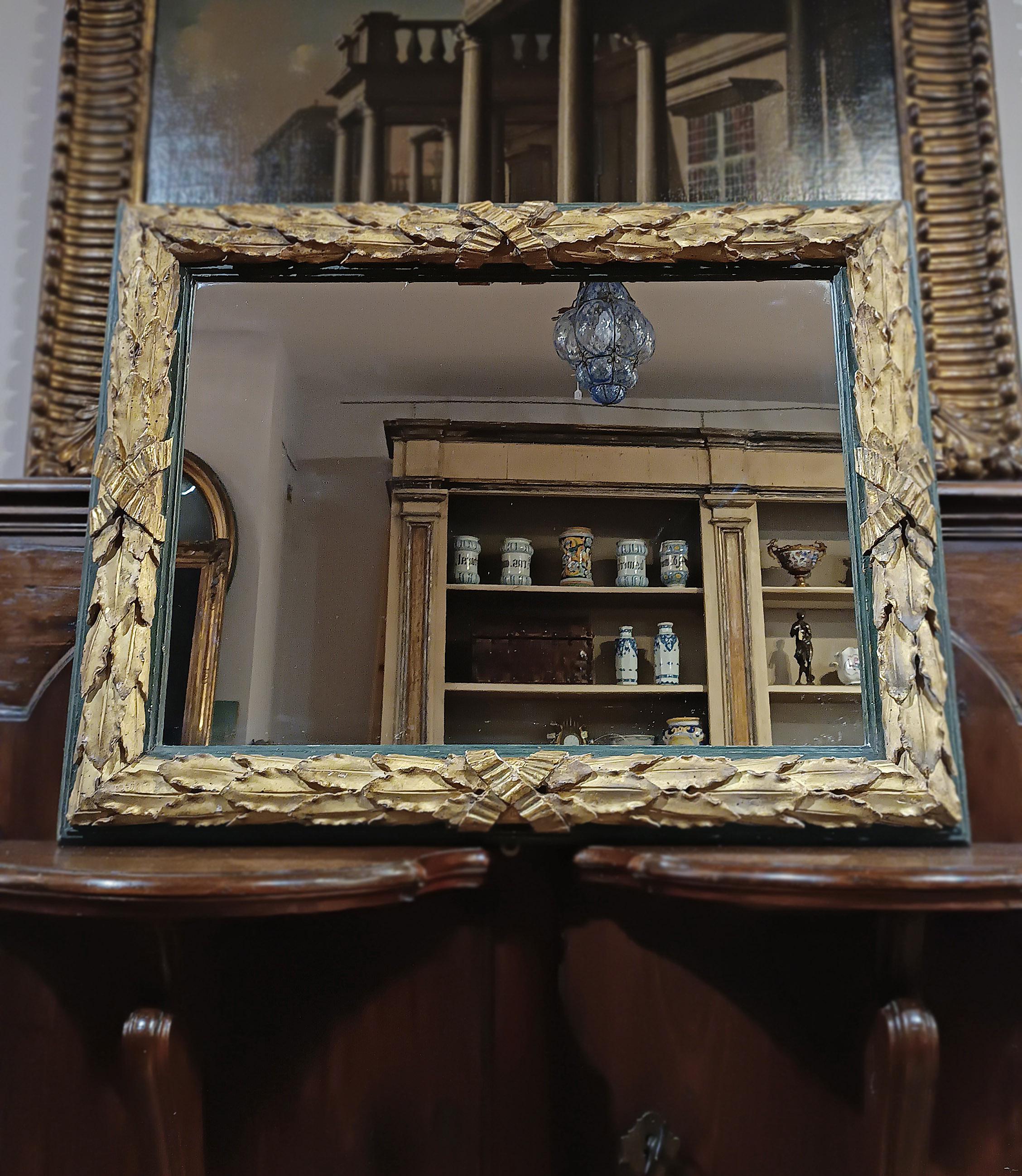 Elegant mirror in carved Swiss pine wood gilded with pure gold leaf, painted in a shade of antique green. The frame is decorated with overlapping laurel leaves and knots at the corners, lending further elegance. The mirror is in good condition and