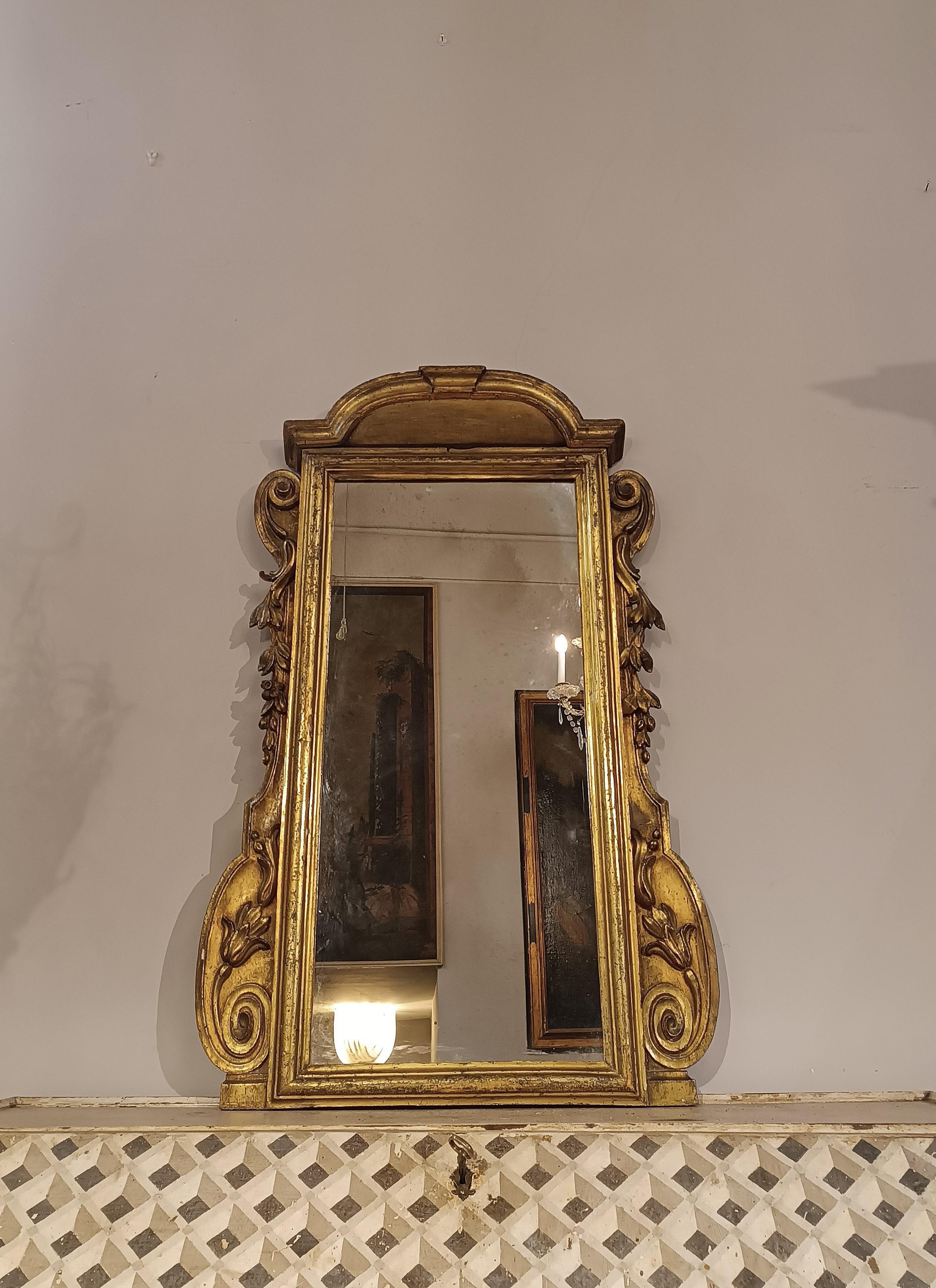 Fascinating small mirror in carved and mecca-gilded wood, characterized by a frame decorated with scrolls and vegetal elements. The cymatium, an architectural substructure and corbel, also adds further charm. Its small size makes it suitable for any