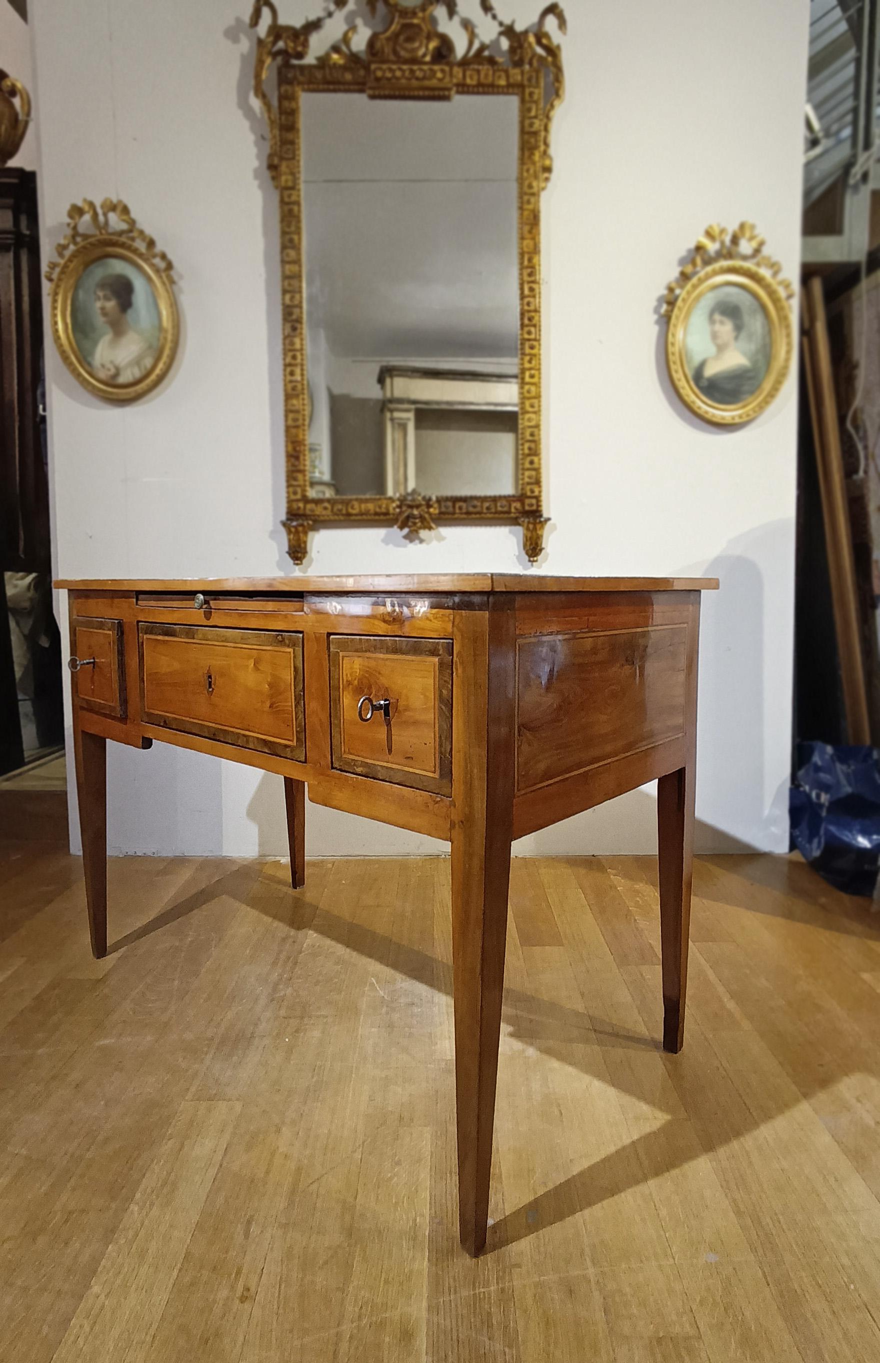 Elegante scrittoio da centrotavola impiallacciato in noce con intarsi di vari legni da frutto. Lo scrittoio si distingue per le sue gambe tronco-piramidali, tipiche dell'epoca, e presenta intarsi geometrici sul fronte, sul piano, sui lati e sul