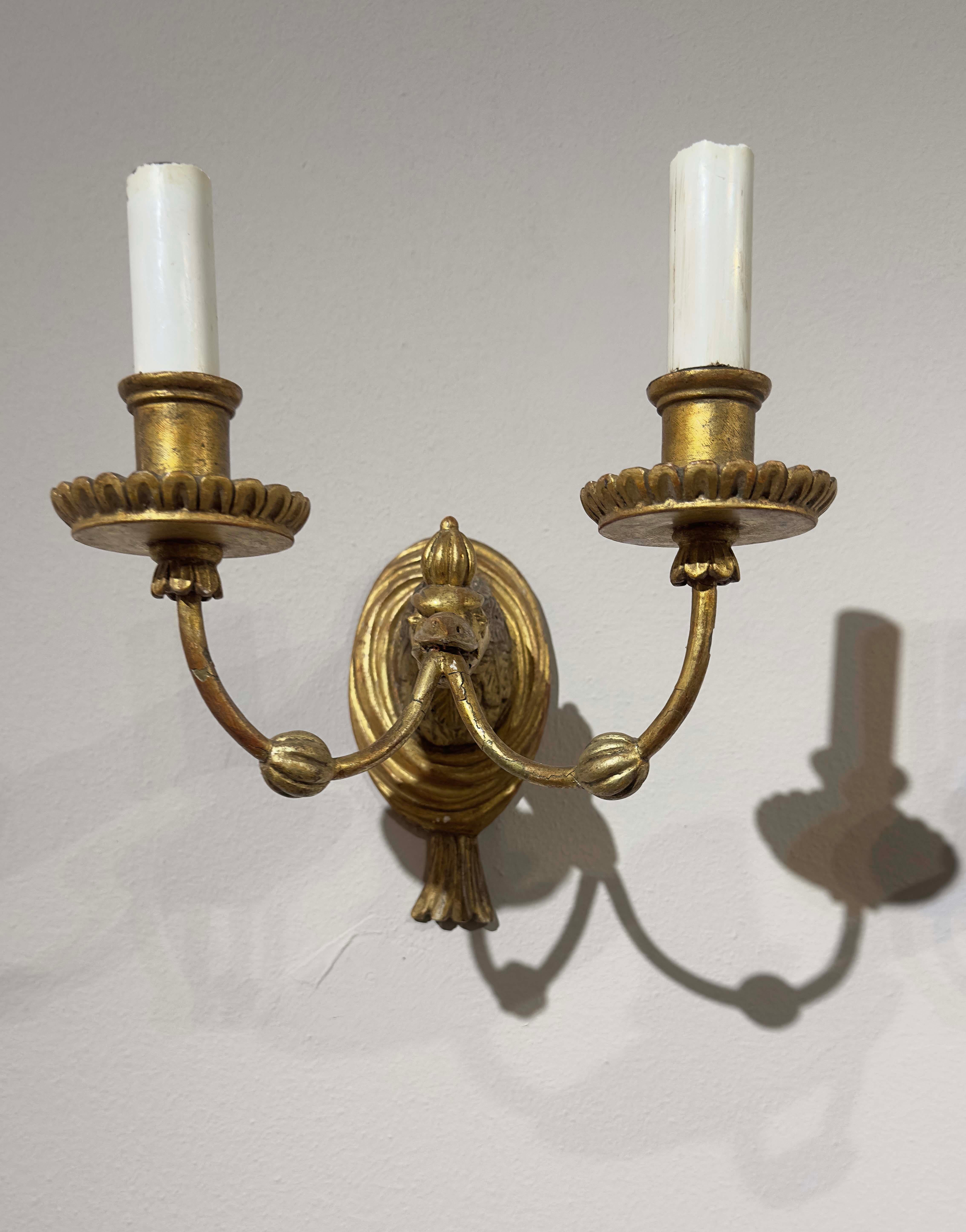 An elegant pair of carved wood wall lights gilded with pure gold leaf. These refined wall lights each have two lights and are electrified. Their distinctive feature lies in the art of carving: the delicate, finely crafted faces of imaginary birds