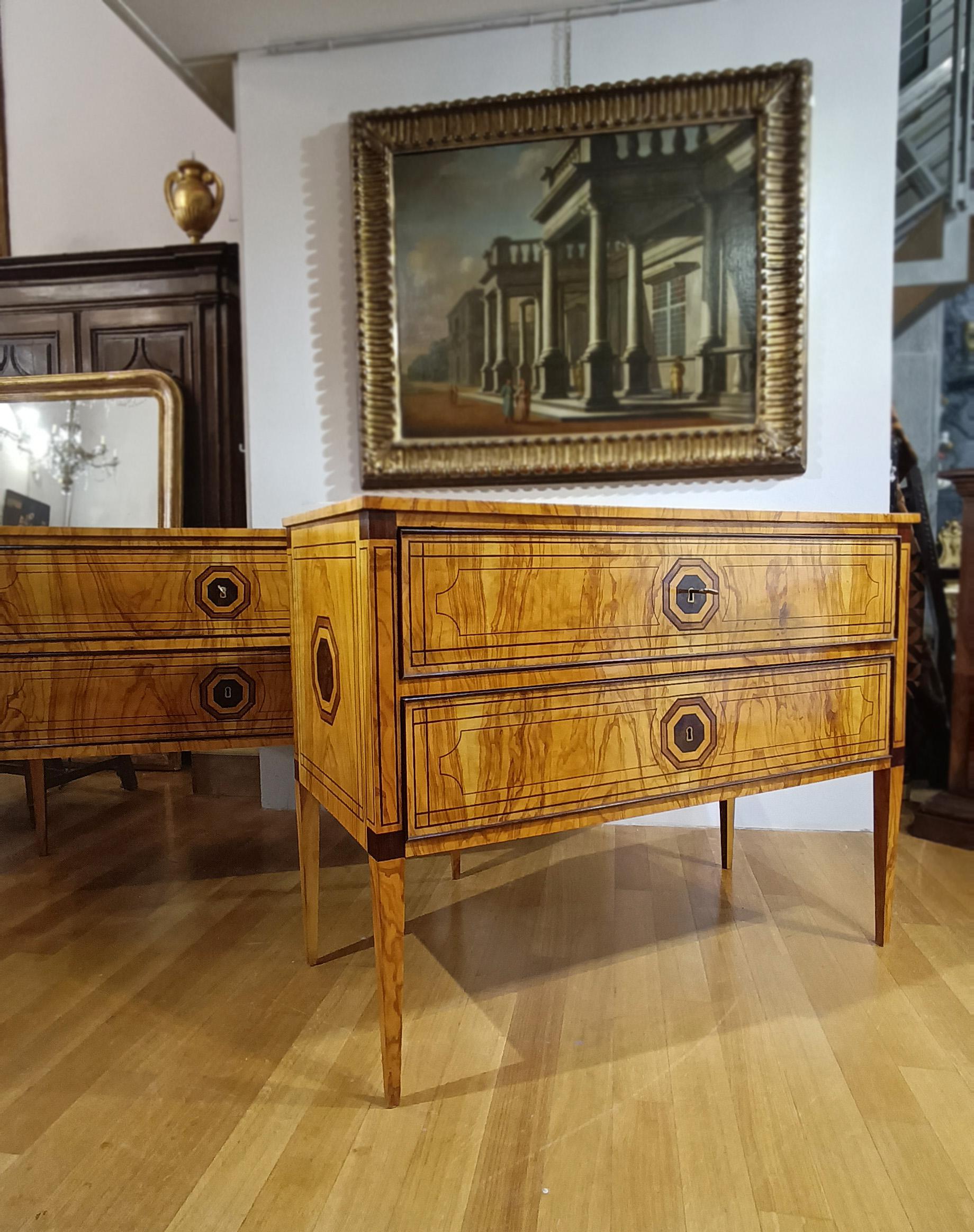 A refined pair of modest-sized chests of drawers, crafted from elm burl veneer with ebonized wood trim. The chests are distinguished by a typically neoclassical style, highlighted by the truncated pyramid legs and the geometric inlay decorations on