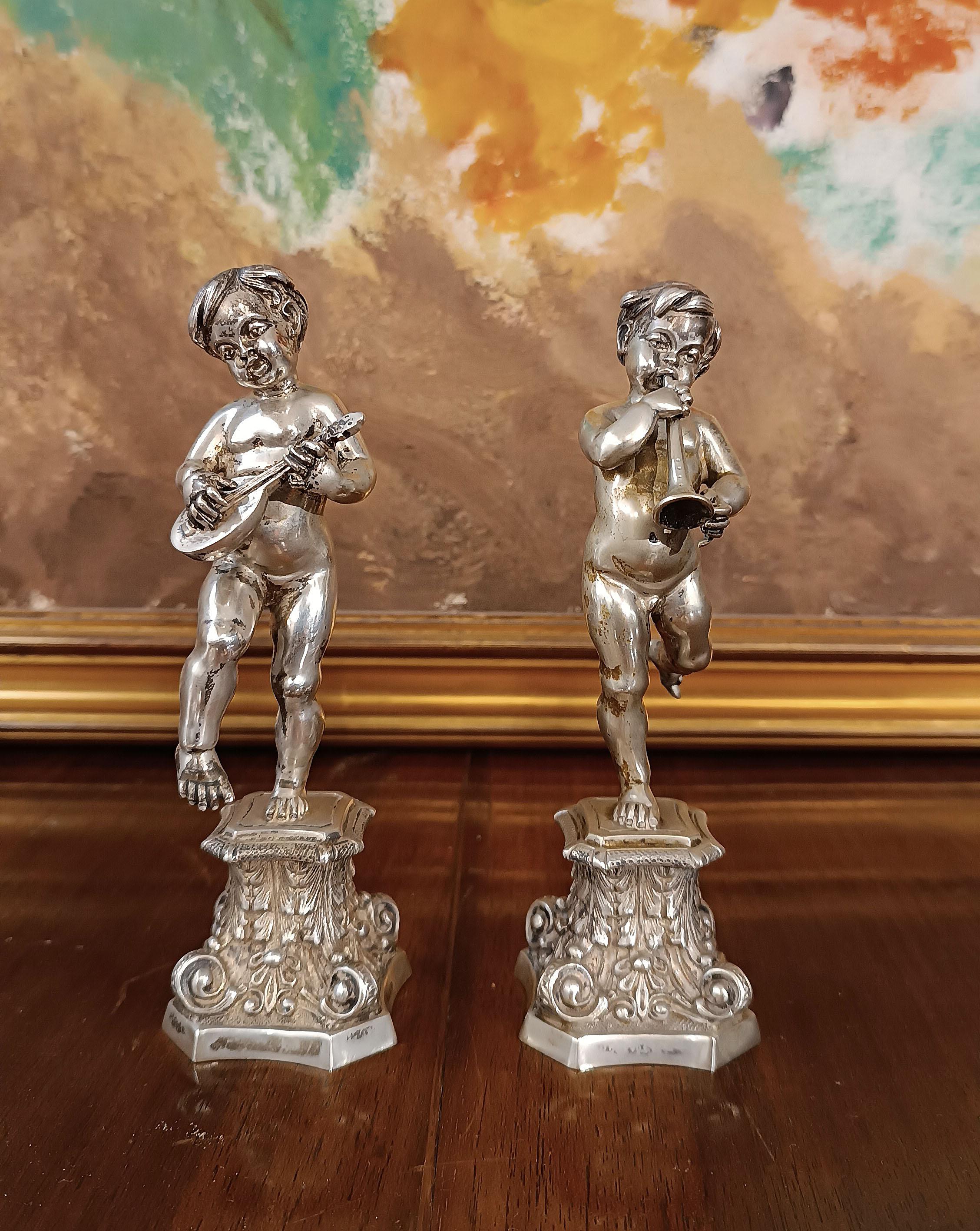 Elegantes Paar silberner Amoretten, die tanzen wollen, während sie ihre jeweiligen Musikinstrumente spielen: der eine mit der Gitarre, der andere mit der Trompete. Die Amoretten stehen auf zwei idealen Sockeln, die durch ein umgekehrtes
