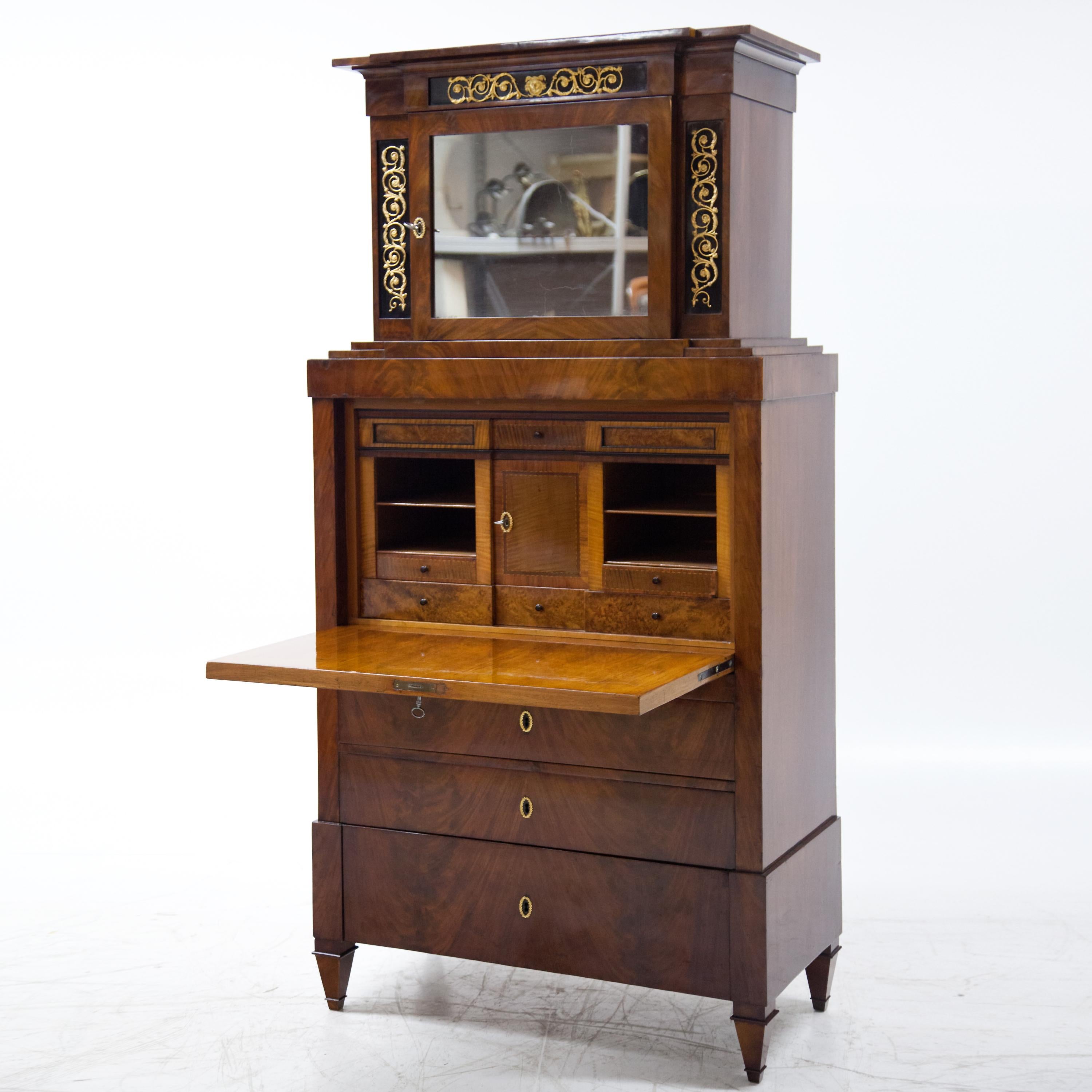 The secrétaire is standing on profiled square tapered feet with a three-drawered chests of drawers base, a writing flap and a one-door top cabinet over a stepped cornice, decorated with fire-gilded vine ornaments and central Hermes mascaron. Inside