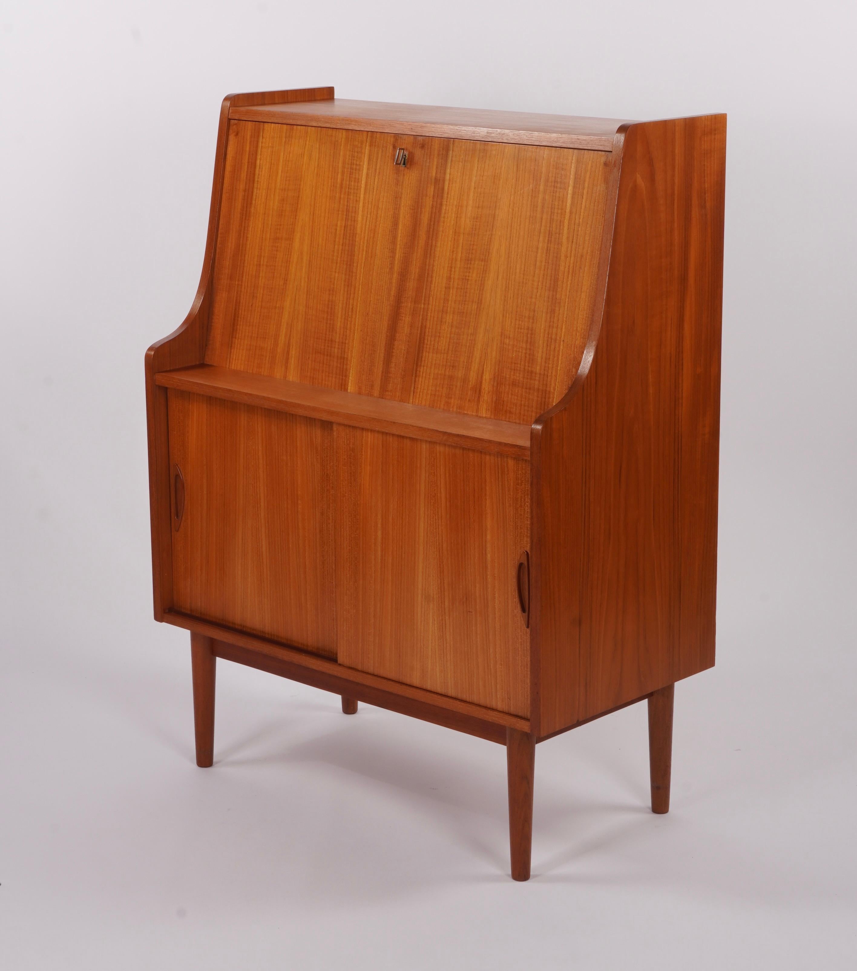 Compact secretary desk by Gunnar Nielsen Tibergaard, constructed of teak and brass. The drop front becomes a desk top surface with four drawers with brass pulls. The bottom half has a storage compartment with two sliding doors with two storage