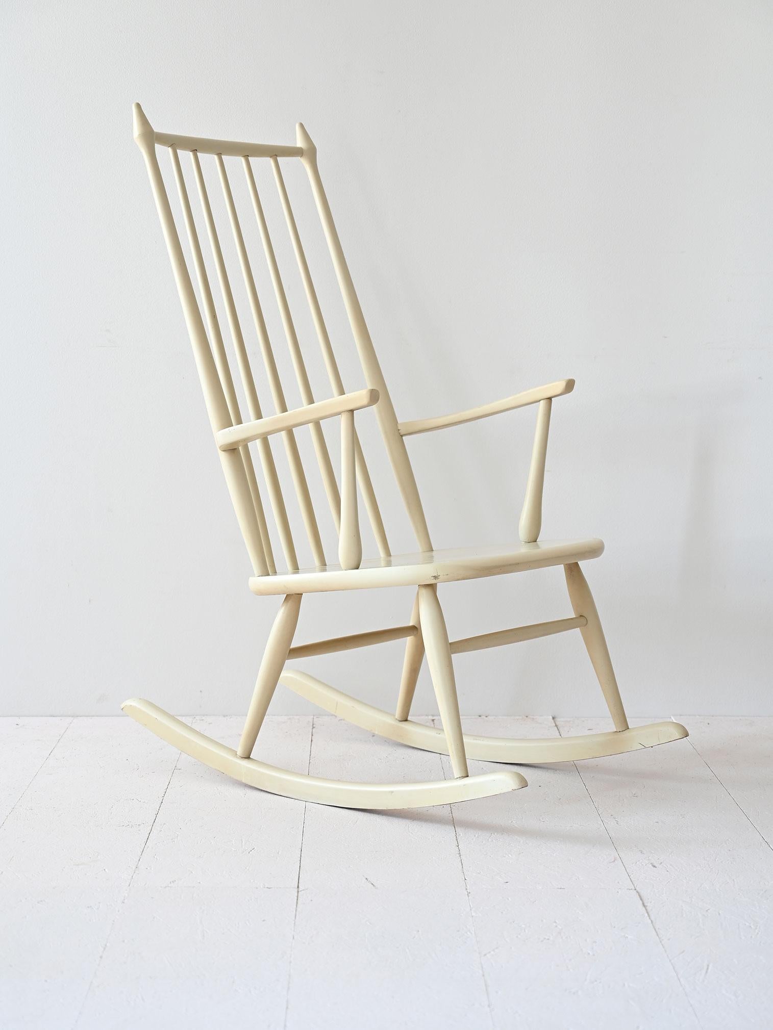 Chaise à bascule scandinave vintage, peinte en blanc, elle se caractérise par un dossier distinctif avec des tiges en bois et un design essentiel et fonctionnel. Idéal pour les environnements lumineux et accueillants, il allie tradition et