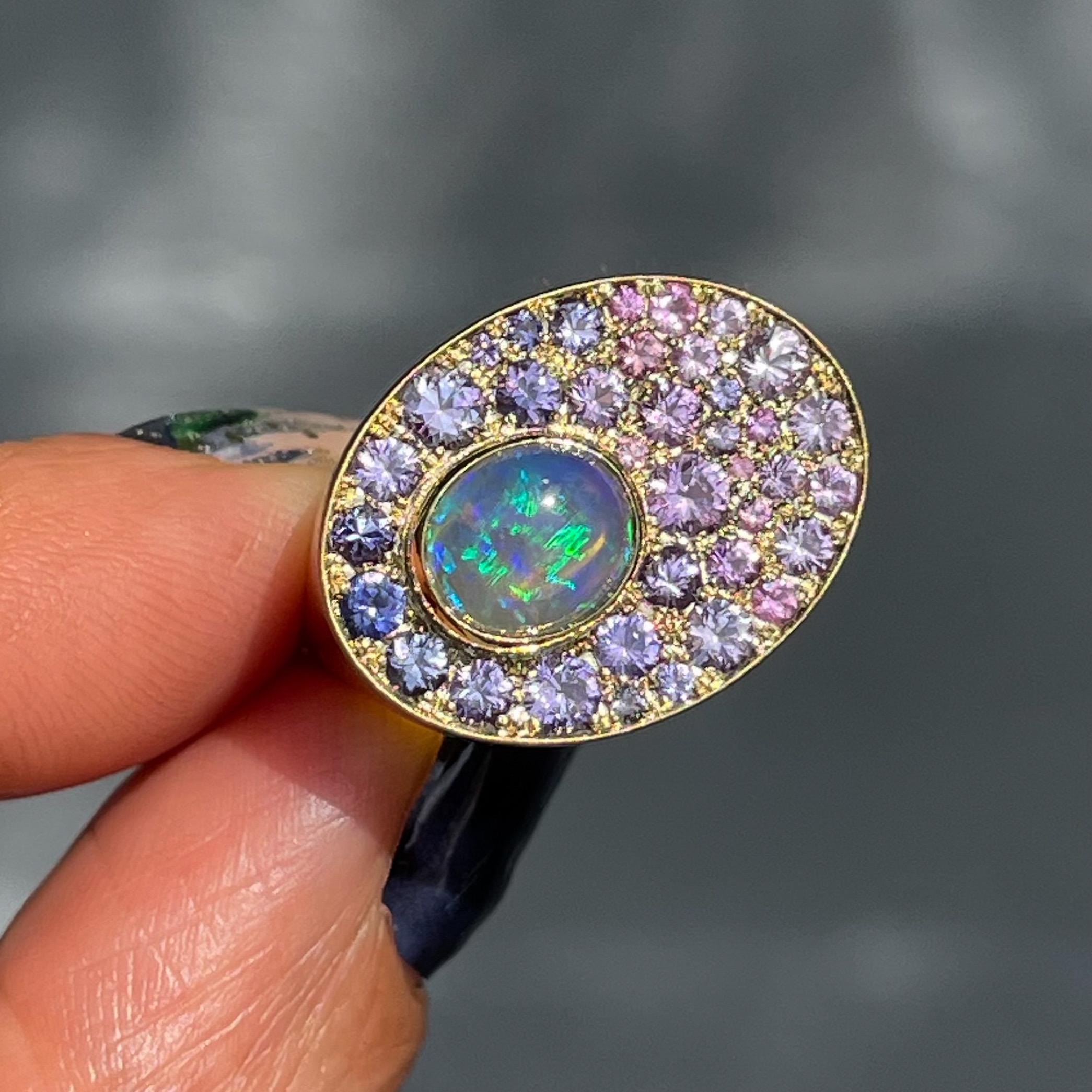 Sur cette bague en opale australienne, une opale cristalline émerge d'un pâturage de saphirs. Comme une couverture de gazon chatoyant, les saphirs roses et violets diffusent leurs teintes autour de l'opale dans un motif ombré fluide. Allant du rose