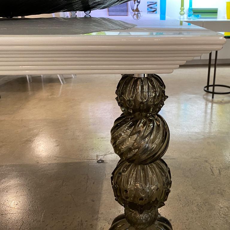 Mori, table carrée avec pieds en verre de Murano par Seguso Vetri d'Arte. Un design élégant et polyvalent qui peut s'intégrer dans un large éventail d'intérieurs. Un plateau laqué blanc est soutenu par 4 pieds en verre de Murano gris. Le style