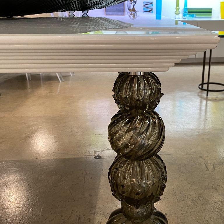 Mori, mesa cuadrada con patas de cristal de Murano de Seguso Vetri d'Arte. Un diseño elegante y versátil que puede encajar en una amplia gama de interiores. La encimera lacada en blanco se apoya en 4 patas de cristal de Murano gris. El estilo
