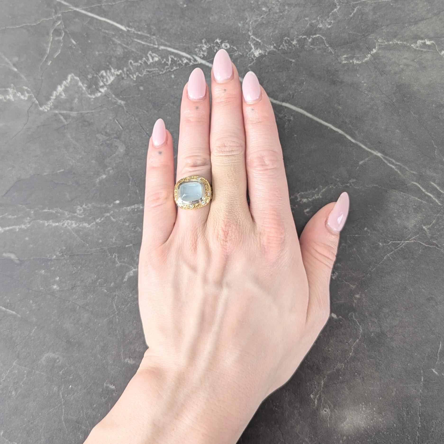 Centering a cushion shaped sugarloaf cabochon measuring 8.5 x 10.5 mm
Transparent milky light blue in color with subtle chatoyancy
Bezel set in white gold with engraved foliate motif surround
Yellow gold and accented by round brilliant cut