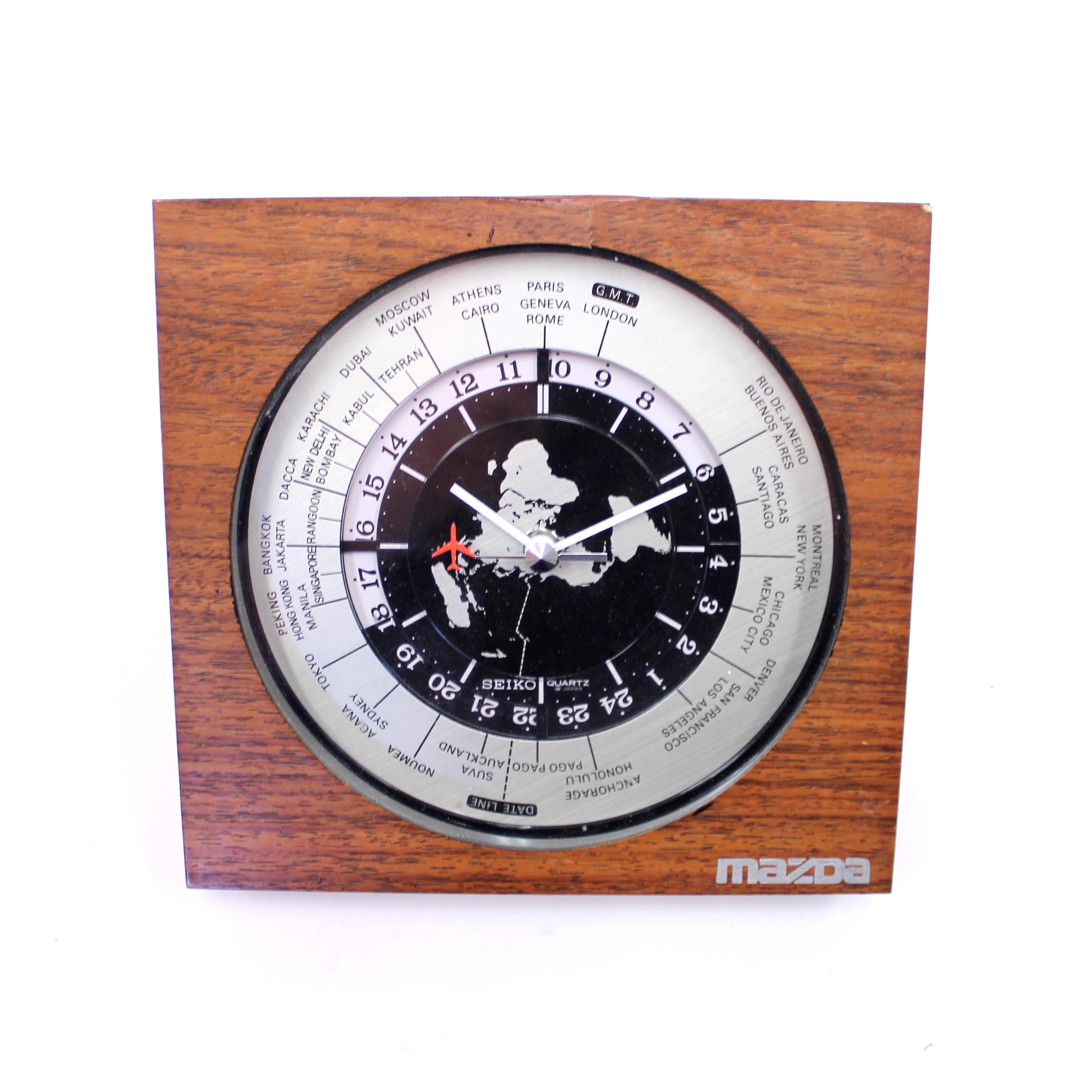 Seiko world timer GMT tilted desk or table clock with a sweeping seconds with a red aeroplane seconds hand and quartz movement from the 1980s. Case made of walnut veneer. This example from a Mazda dealership. Good untouched vintage condition with a