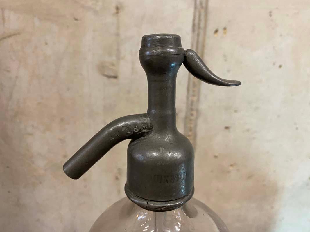 Antique siphon from France from the years around 1900. The bottle is made of thick heavy clear glass and has a closure made of tin. In the cap is stamped the word UINETTE and the sequence of numbers 10165. The glass body has the words 