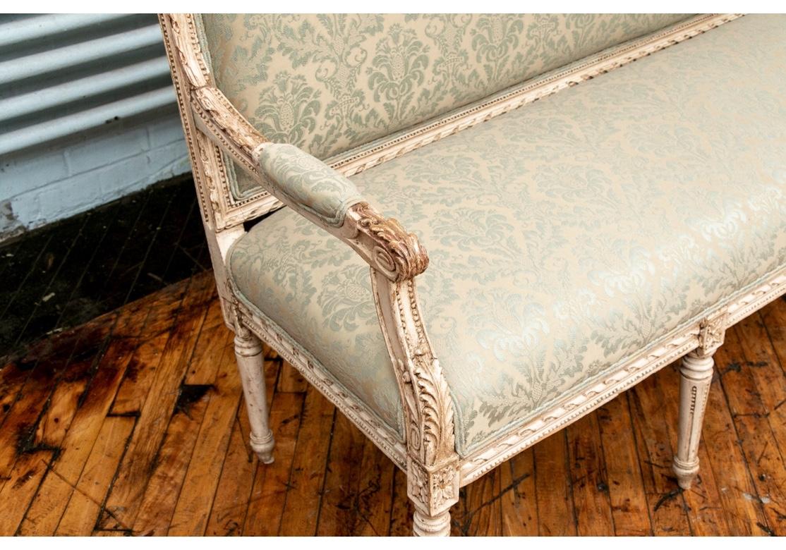 Classic Louis XVI style salon sofa carved and painted in ecru. With a ribbon and beaded carved frame overall, the shaped crest rail with floral carved crest. The arms with carved acanthus leaves on the ends and curved supports. Raised on four