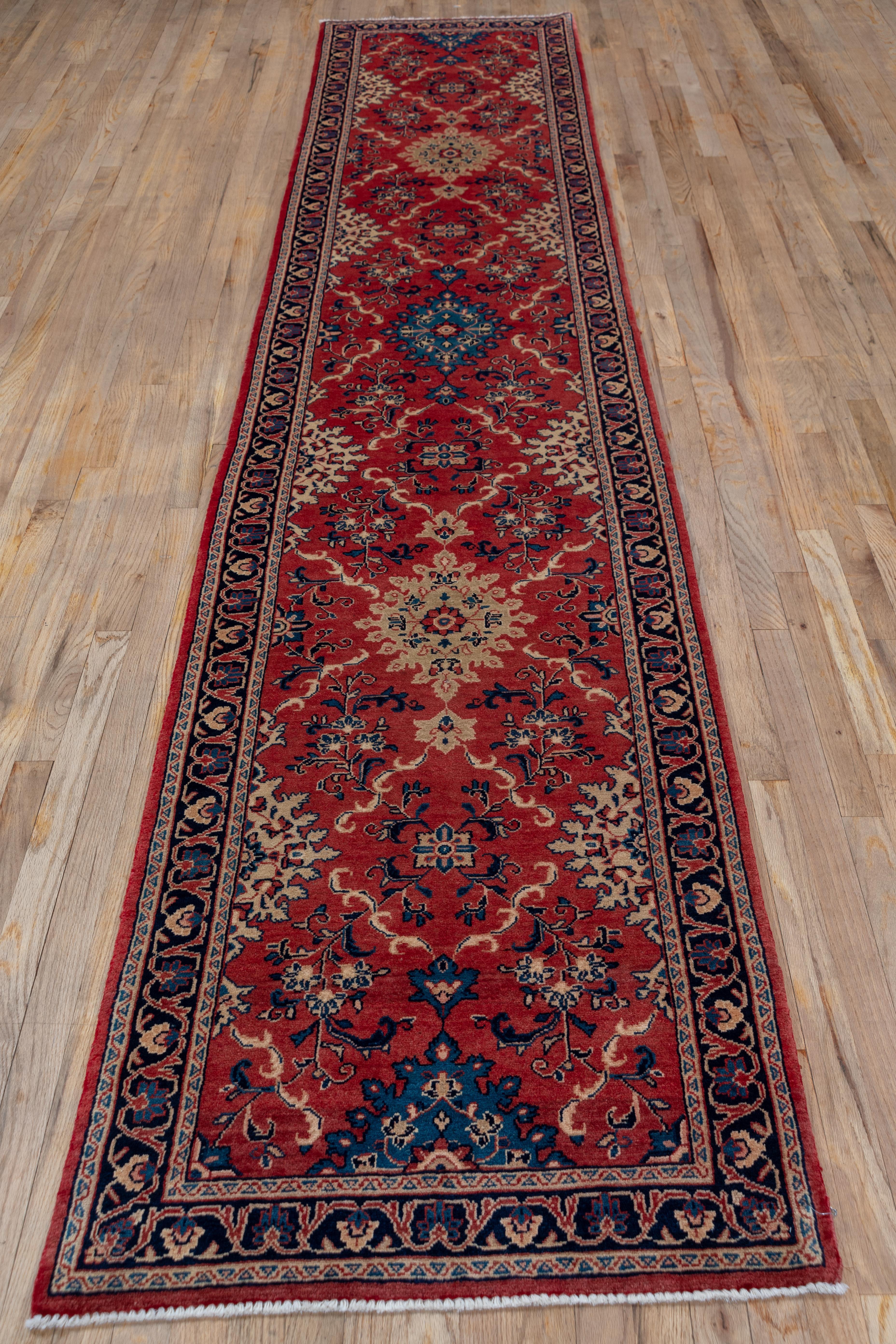 The red ground is decorated by long flowering tendrils, leafy lozenges and small octogrammes, all in sand, navy, beige and teal. The navy border fits palmettes into a split arabesque pattern. The condition is excellent as befits this semi-antique