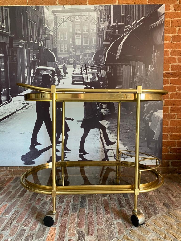 This 1980s vintage Italian Regency style oval bar cart has a brass frame with smoked glass. The brass frame features two levels. The top level features a smoked glass shelf with a bar around the top with a handle to push the cart. The bottom shelf