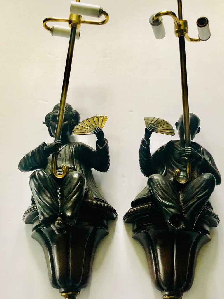 A fabulous and rare pair of heavy bronze wall sconces by Maitland Smith. In the Asian style, these wall sconces depict a seated man and a seated woman holding fans and umbrellas in brass. The bases are made to look like distressed wood and have