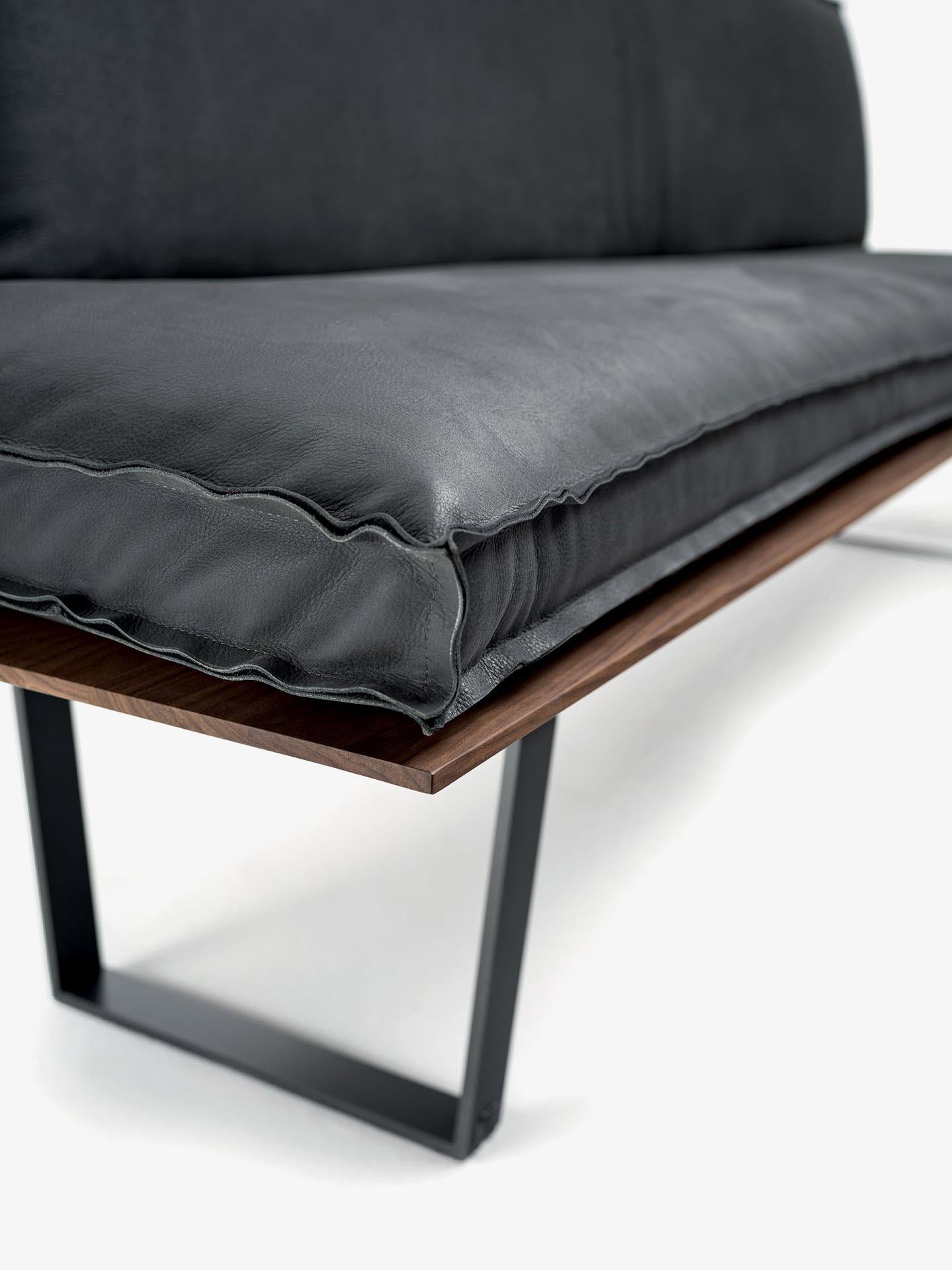 Banc avec assise en bois massif en listes collées et pieds en fer. L'assise et le dossier sont équipés de maxi-coussins recouverts de cuir ou de tissu avec surpiqûres apparentes, en deux versions différentes. Disponible en version linéaire et en