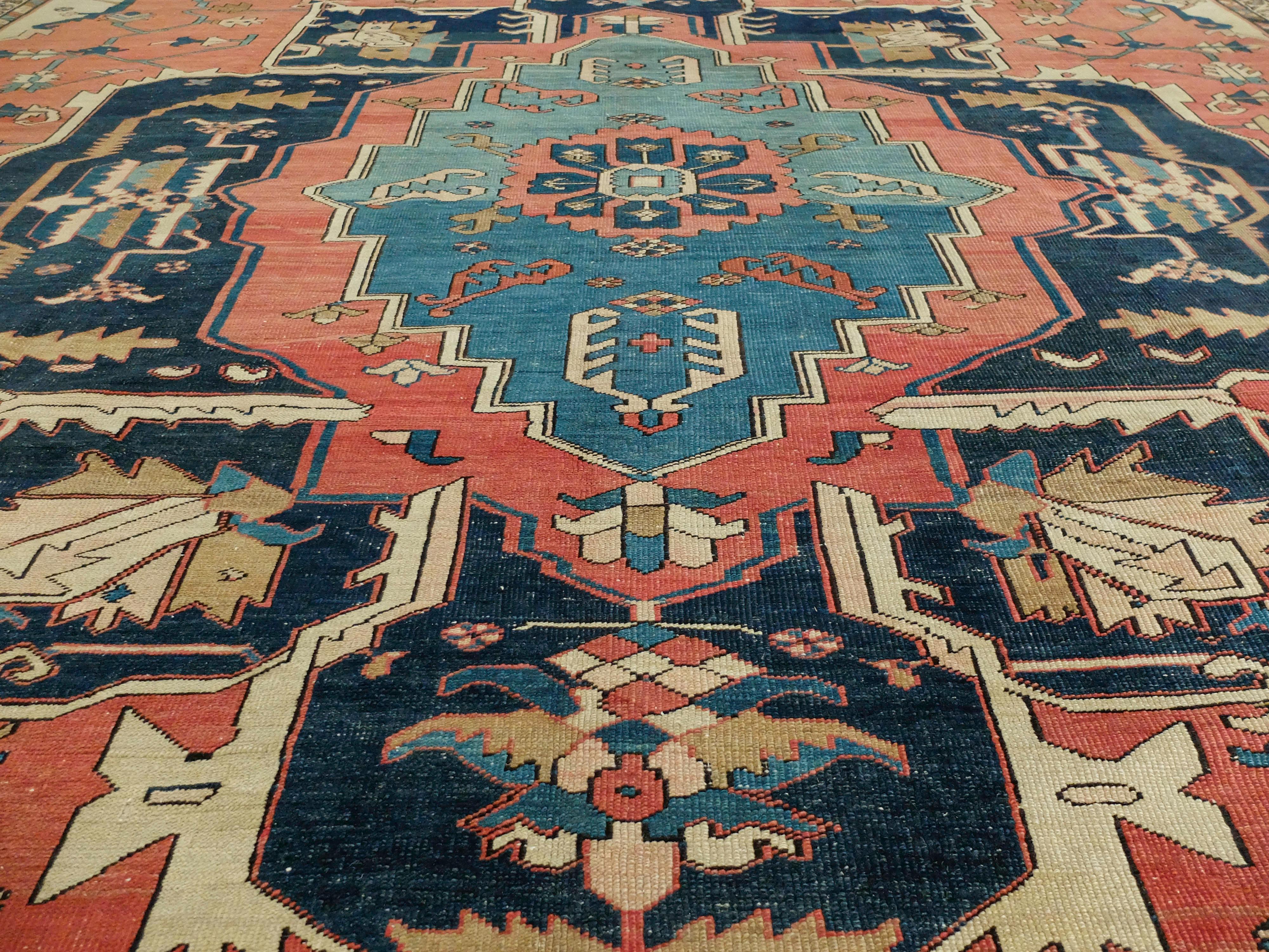 This is a Serapi rug made circa 1880s. It has a central floral like medallion that is surrounded by a soft reddish terra cotta color. It also has 4 structures in the corners before the border patterns begin. The border patterns are more dense and