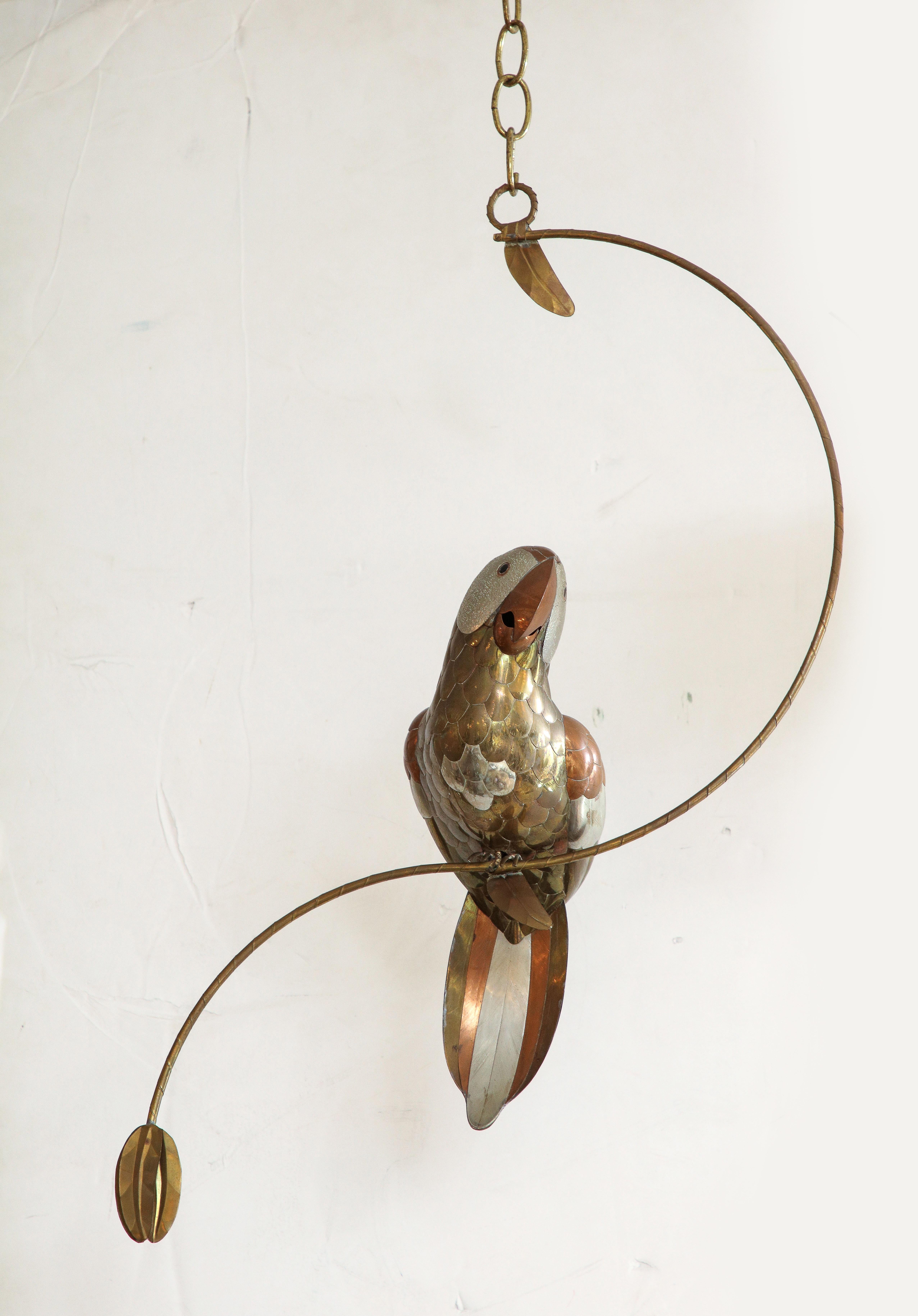 Modernist mixed metal handmade Parrot sculpture composed of brass, copper and tin sitting on a stylized tree limb.Overall height with chain is 36, inches,  parrot with limb measures 25