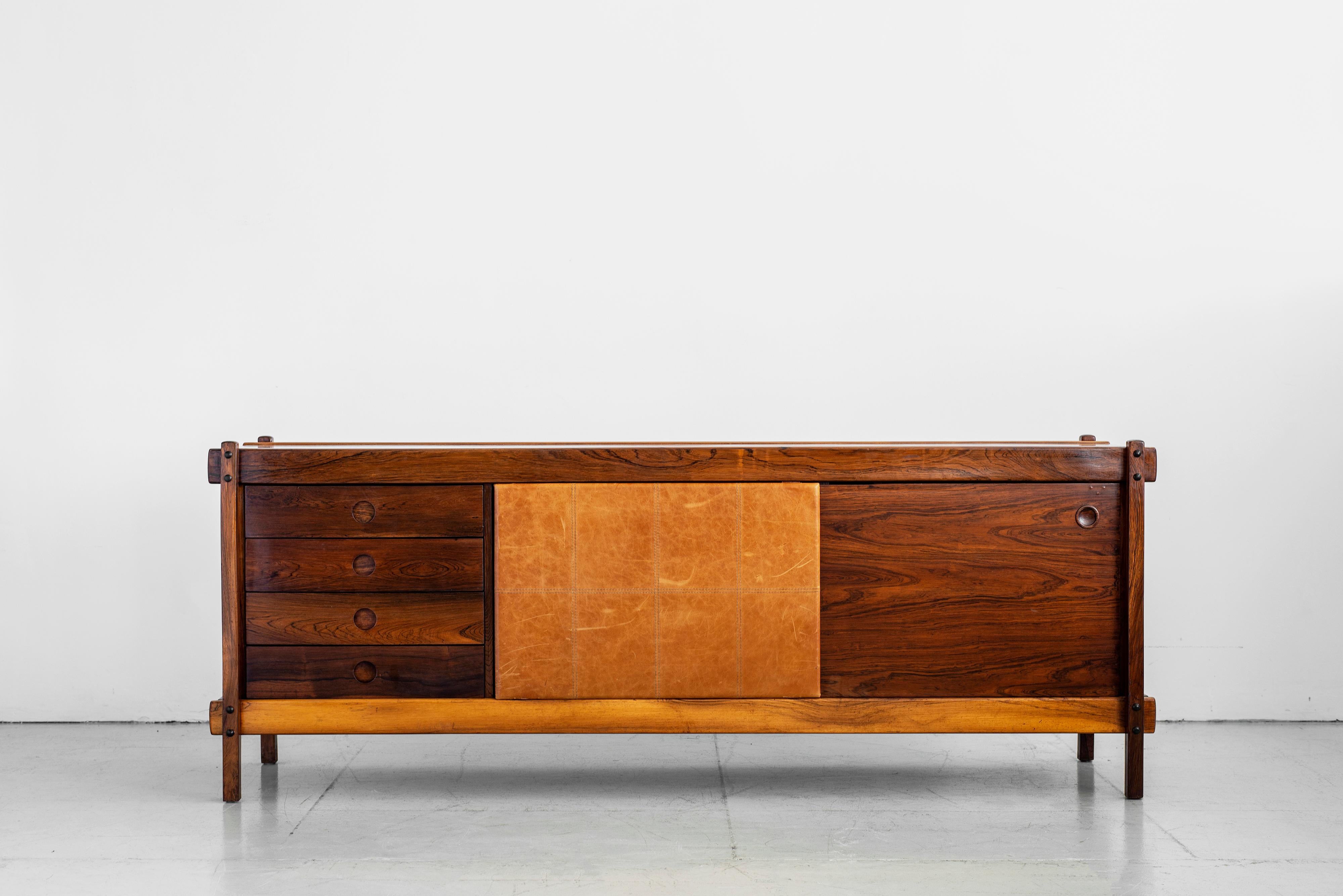 Handsome Sergio Rodriguez sideboard made of beautiful rosewood with stunning grain. Central panel door in brown saddle-stitched leather slides to reveal open shelvings but with exposed 4 drawers on the other. Original labels on back. Incredible