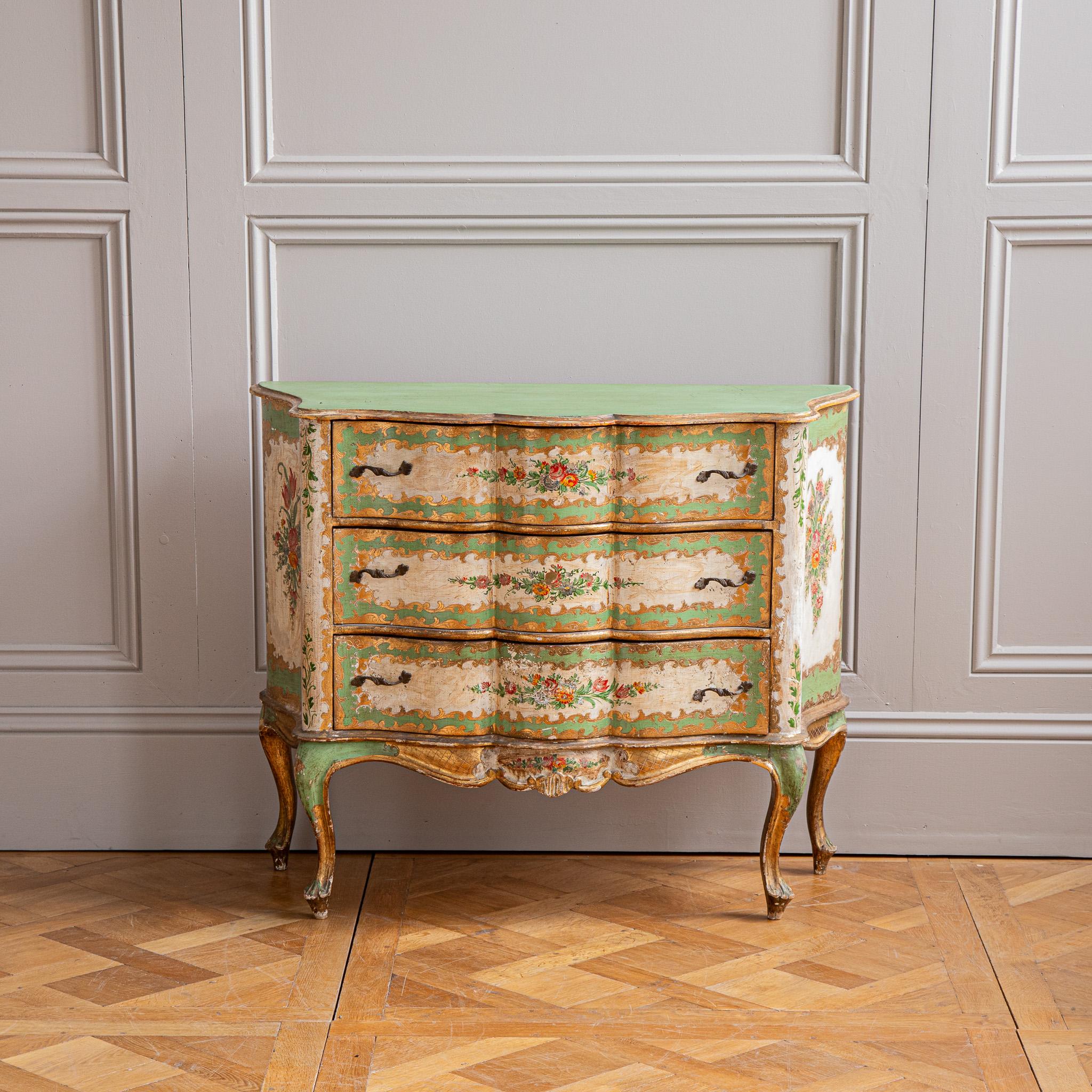 Cómoda italiana rococó pintada, con frente de serpentina, provista de tres cajones, el frente y los laterales están decorados con motivos florales sobre fondo verde y dorado .  La cómoda se alza sobre patas cabriolé y tiene tiradores de latón.