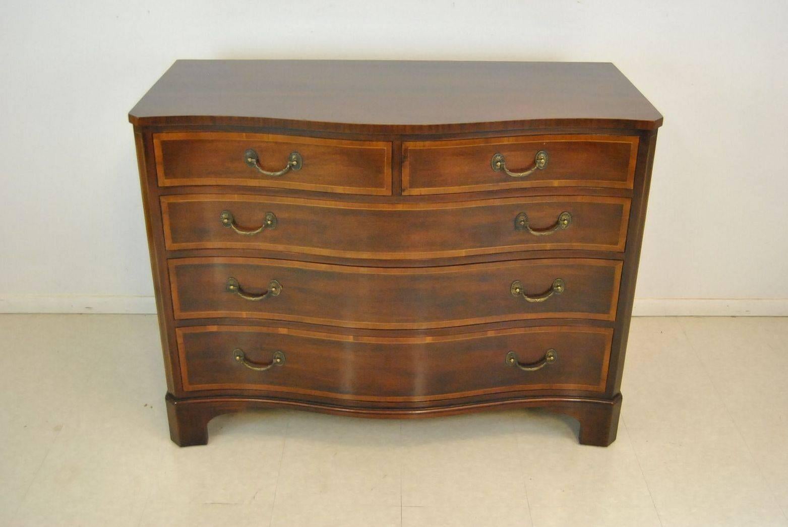 A serpentine front mahogany chest/dresser made by Kindel Furniture. This chest has a nice hand done inlay pattern on the sides. The drawers have the same fine line inlay with a larger border done in a different tone. The chest has its original brass