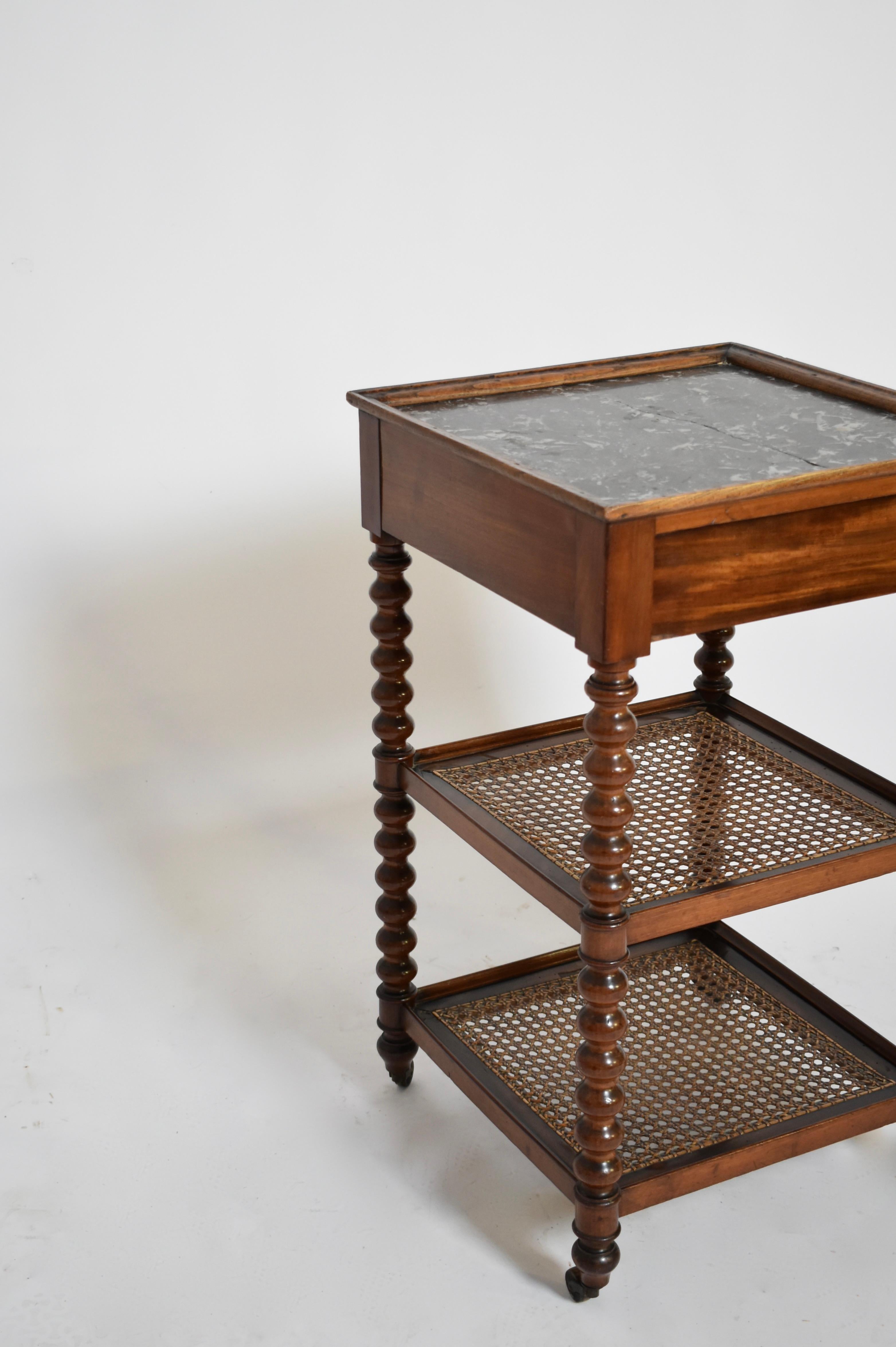 Tavolino o comodino composto da quattro piedi torniti che terminano con rotelle in bronzo che sostengono due vassoi di canna e un piano in marmo nero con un cassetto nella cintura. Inghilterra, fine del XIX secolo.