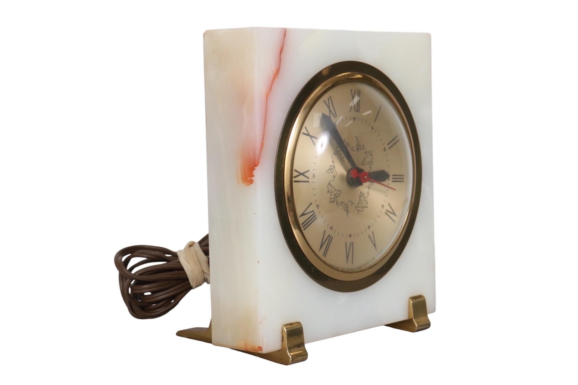 A mid century electric mantle clock. The clock face is decorated with a vine leaf motif and clover details, set in a marble block with a crimson vein in the top corner. Rests on brass feet.
   