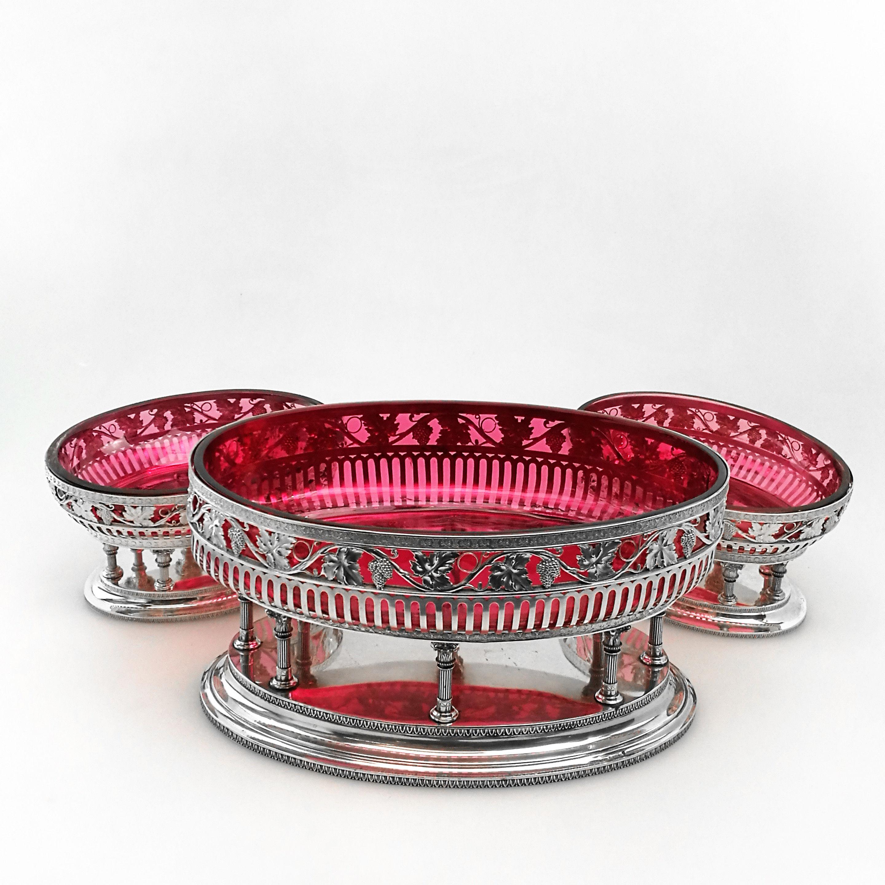 A set of three Antique solid Silver Comports with deep red claret colored glass liners. The set consists of one large oval shaped dish and two matched smaller oval dishes. Each dish has a solid oval pedestal foot and the dishes rest on a series of