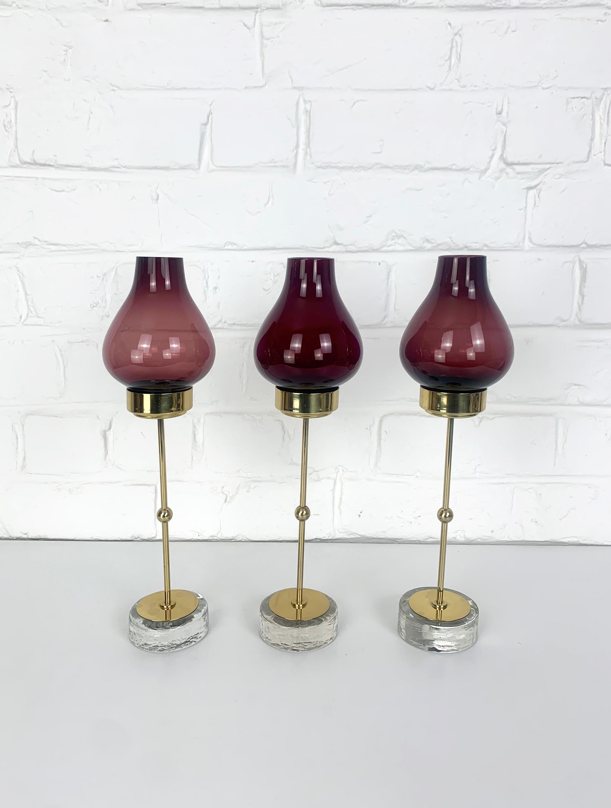 Set of 3 Swedish Modernist candle holders by Gunnar Ander. Produced by Ystad-Metall, located in the town of Ystad in Sweden. 

Candle-holders in solid, polished brass on a base of clear glass with an irregular surface pattern. Glas shades in