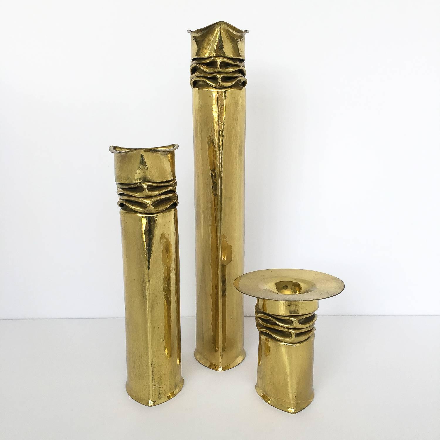Set of three Thomas Roy Markusen Brutalist brass candlesticks. These candle holders or vases feature a brass-plated hammered triangular metal form with crumbled detail. Wide round flat flange on smallest example. Stamped on the bottom of each - TRM