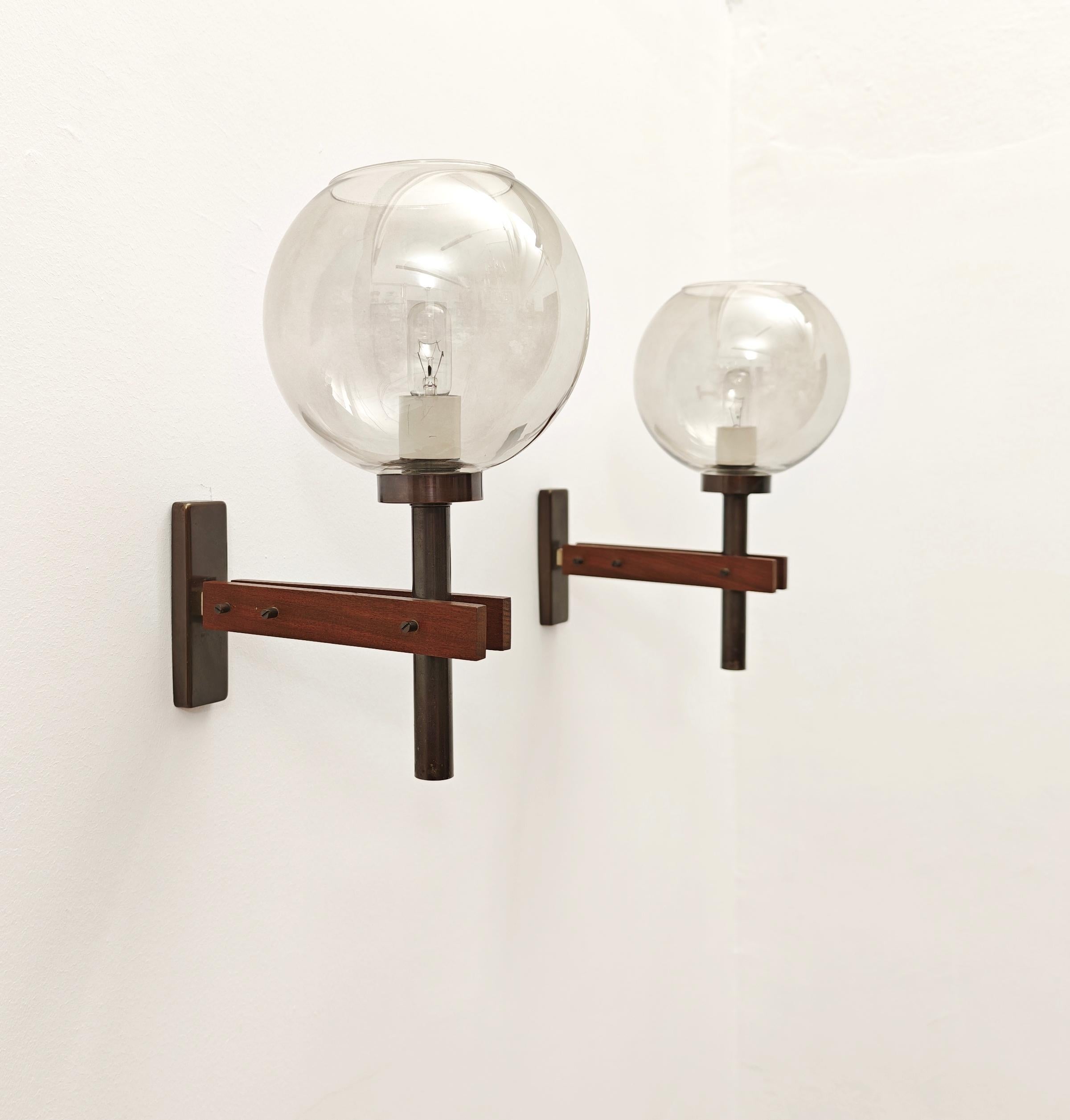 Dos apliques con estructura de latón bronceado y madera, difusor esférico de cristal transparente en tonos ahumados. Placa mural rectangular de latón bronceado. Atribuido al diseño de Frattini. Producción italiana, aprox. años 60. Observa