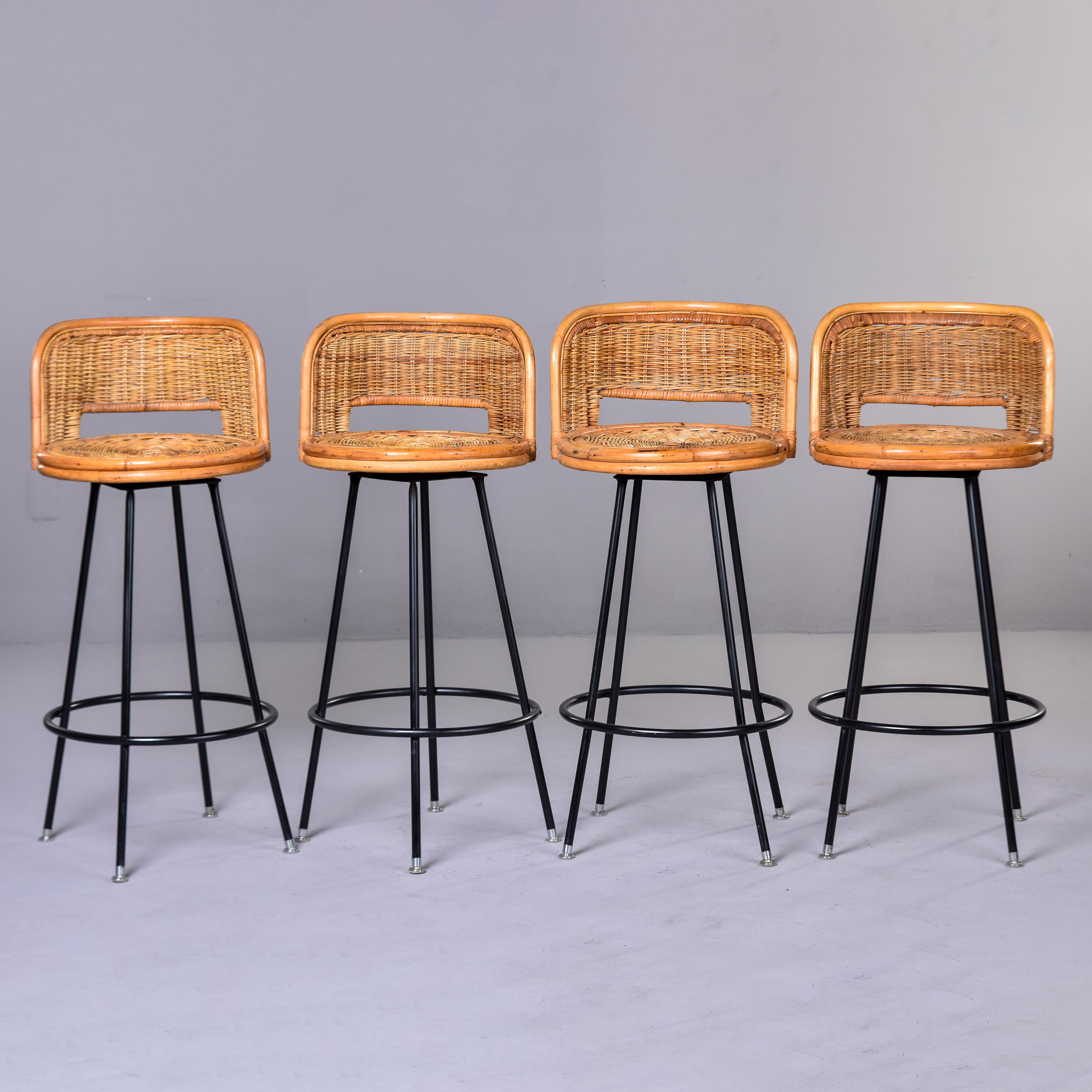 Found in the U.S., this set of four woven wicker swivel bar height stools date from the early 1960s. Stools have a black finished metal base with silver tone metal foot caps, round seats, and low back rests in the style of Danny Ho Fong. Very good