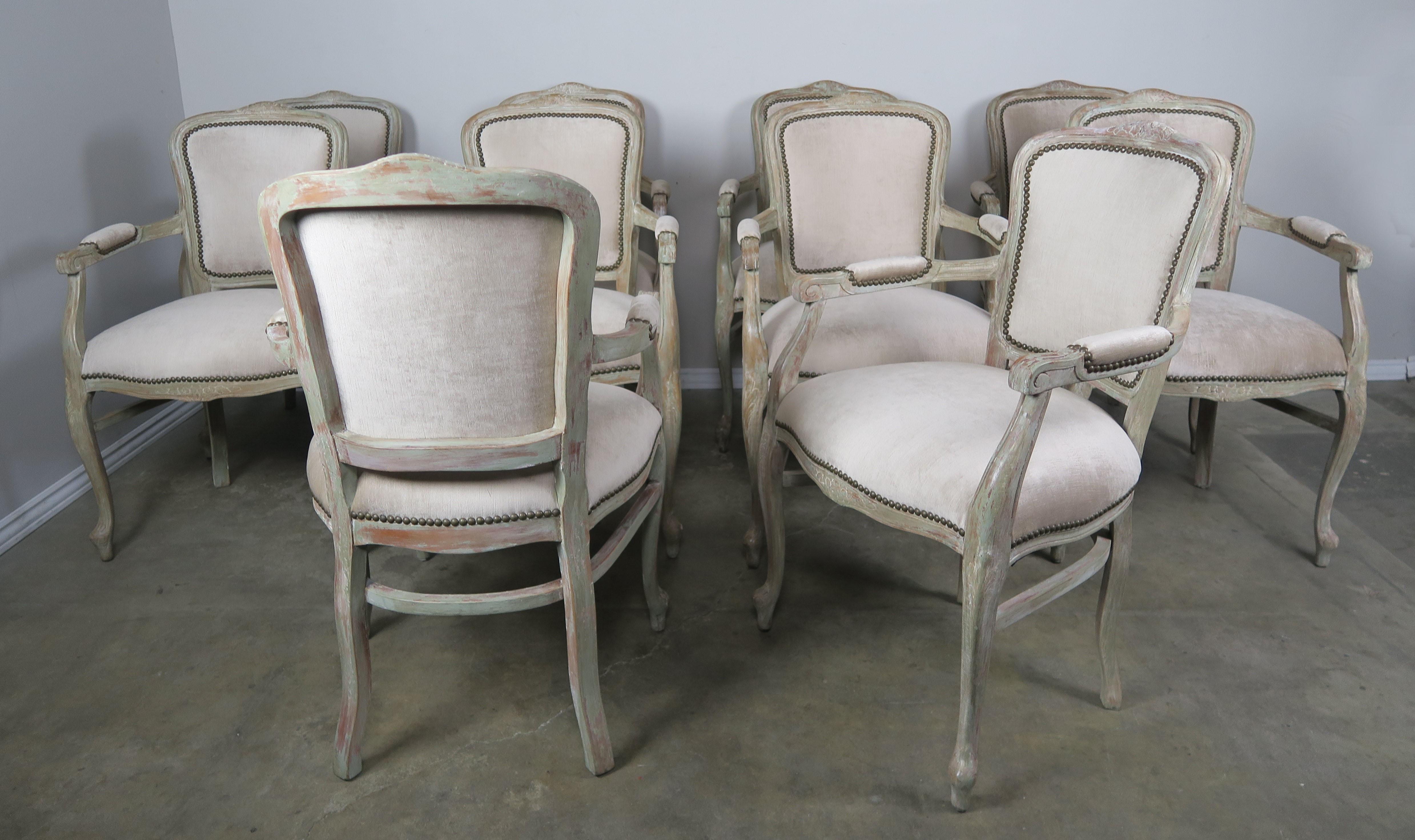 Set of ten French carved Louis XV style armchairs softly painted with remnants of aqua paint left behind. The armchairs are newly upholstered in a cream colored linen velvet with antique brass nailhead trim detail. The chairs stand on four cabriole