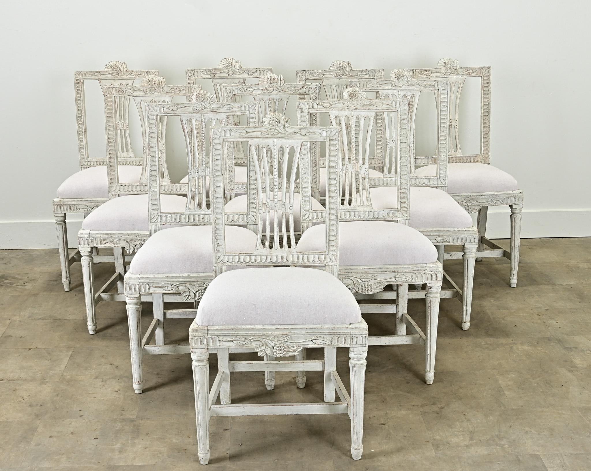 A stately set of ten Swedish Gustavian upholstered dining chairs from the Louis XVI period. These sturdy and comfortable painted chairs are all hand carved with a floral and hop design at the top of the chair back and leaf motifs on the other three