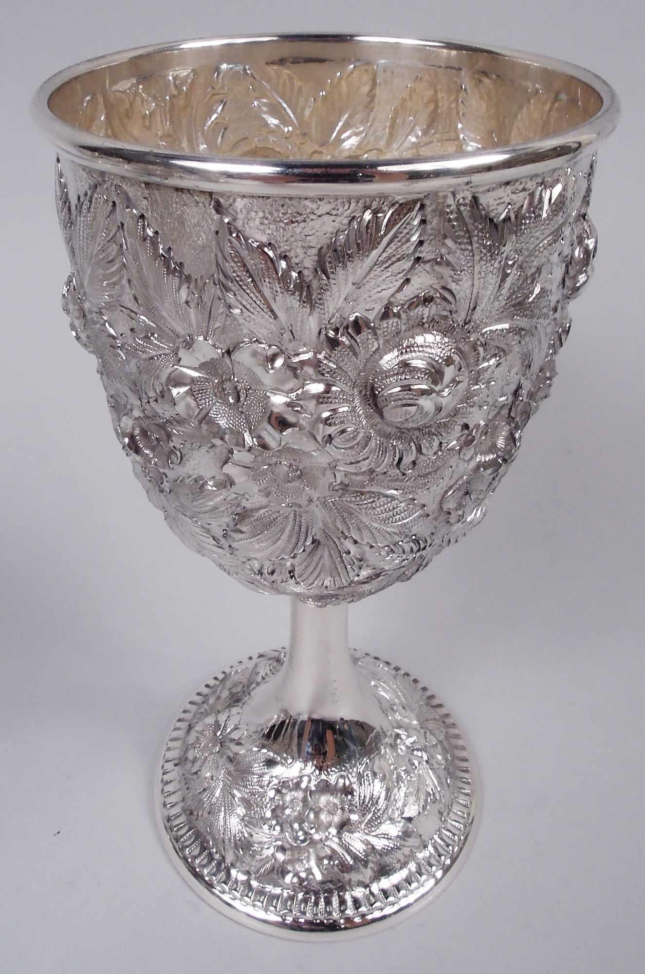 Set of 12 Edwardian sterling silver goblets. Made by S. Kirk & Son Inc. in Baltimore. Each: Ovoid bowl on cylindrical stemp flowing into domed foot. Dense fronds and flowers repousse. Mouth rim molded and foot rim crimped. Tactile easy-grip form