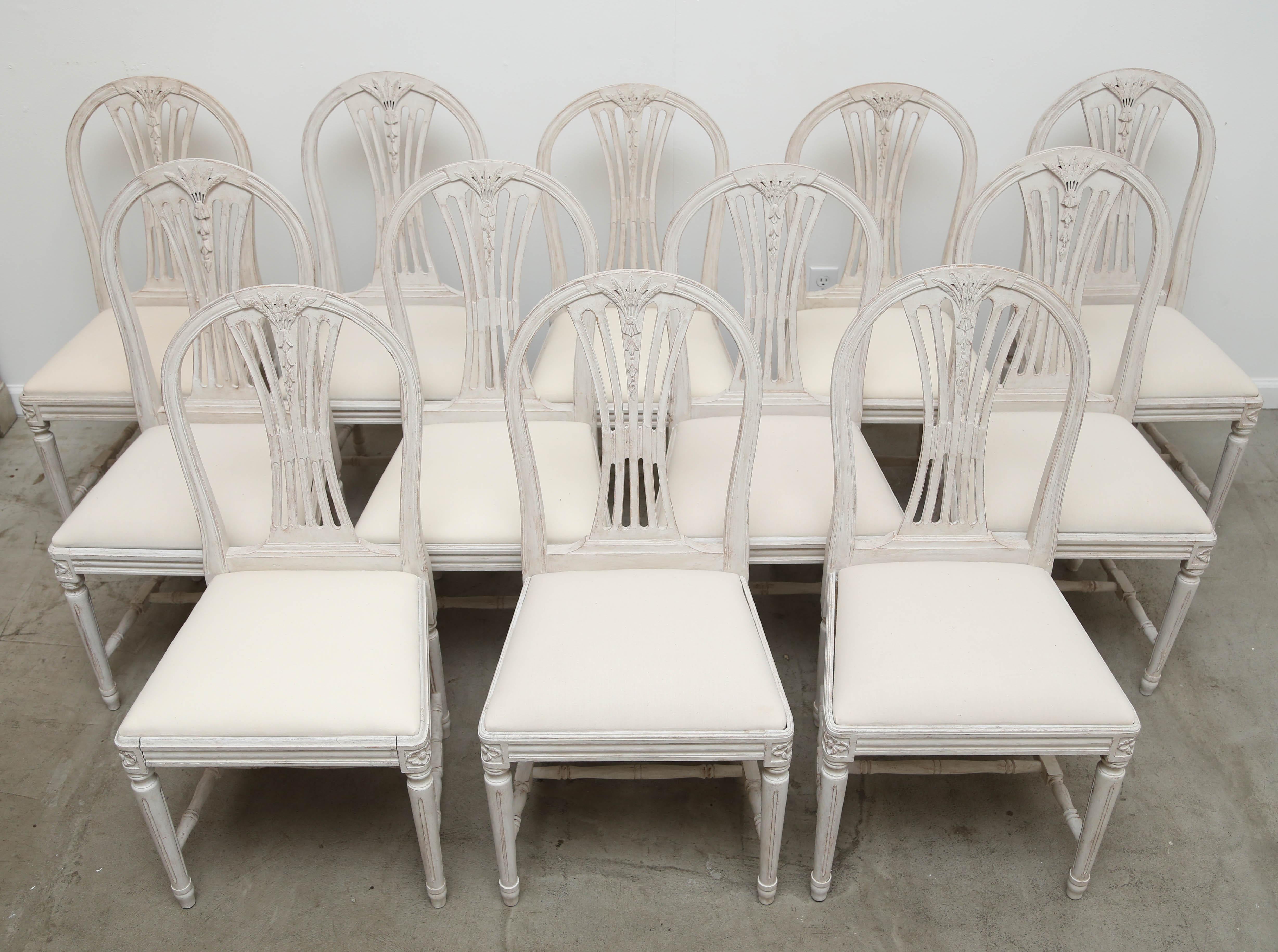 Set of 12 antique Swedish painted Gustavian style dining chairs, each with arched backs carved with wheat and bell flowers, openwork spelt, square drop-in seats, round fluted legs, joined by H-shaped stretchers, and carved rosettes on top of seat