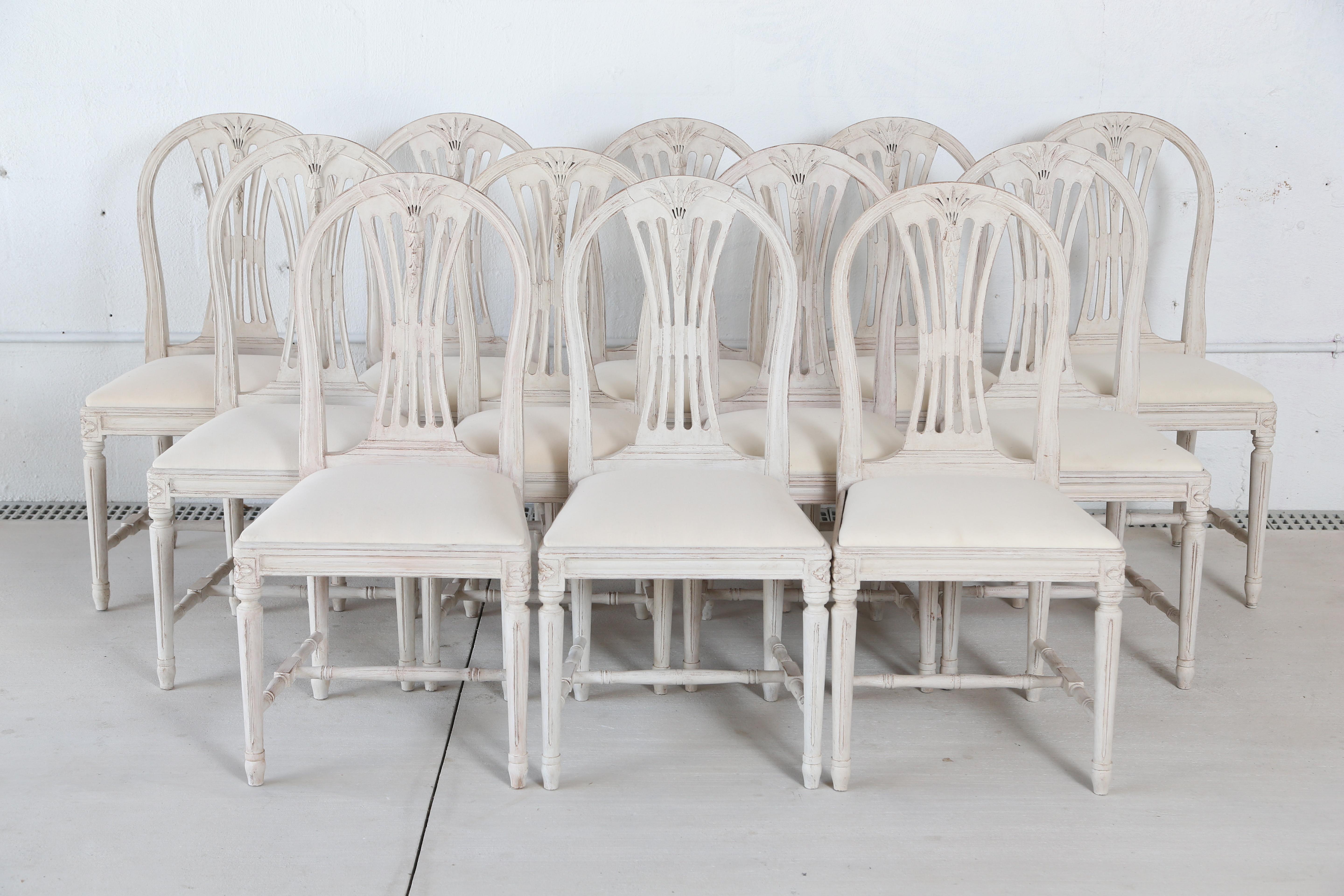 Set of 12 antique Swedish painted Gustavian style dining chairs, each with arched backs carved with wheat and bell flowers, openwork spelt, square drop-in seats, round fluted legs, joined by H-shaped stretchers, and carved rosettes on top of seat
