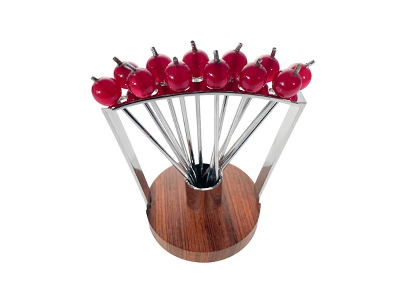 12 chromed cocktail picks with forked points and red 'cherry' tops arranged in two rows of 6 in an arched chrome frame rising from a wood disk base with a central chrome cylinder which the picks hang in.
