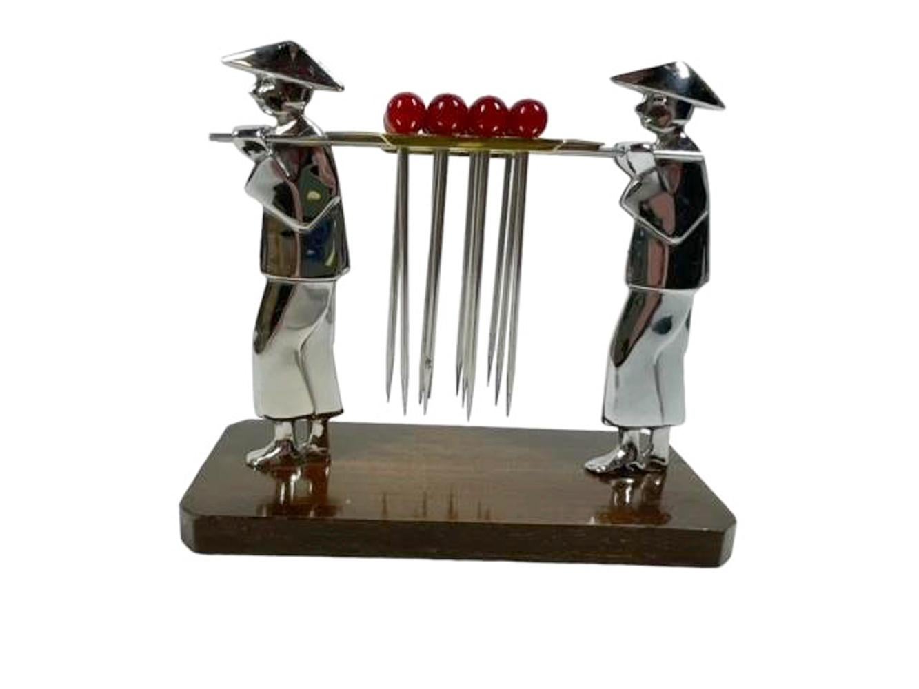 Set of 12 cocktail picks with red Bakelite ball tops and forked picks resting on a gold-tone pallet carried by poles on the shoulders of two chrome finished men standing on a wood base.