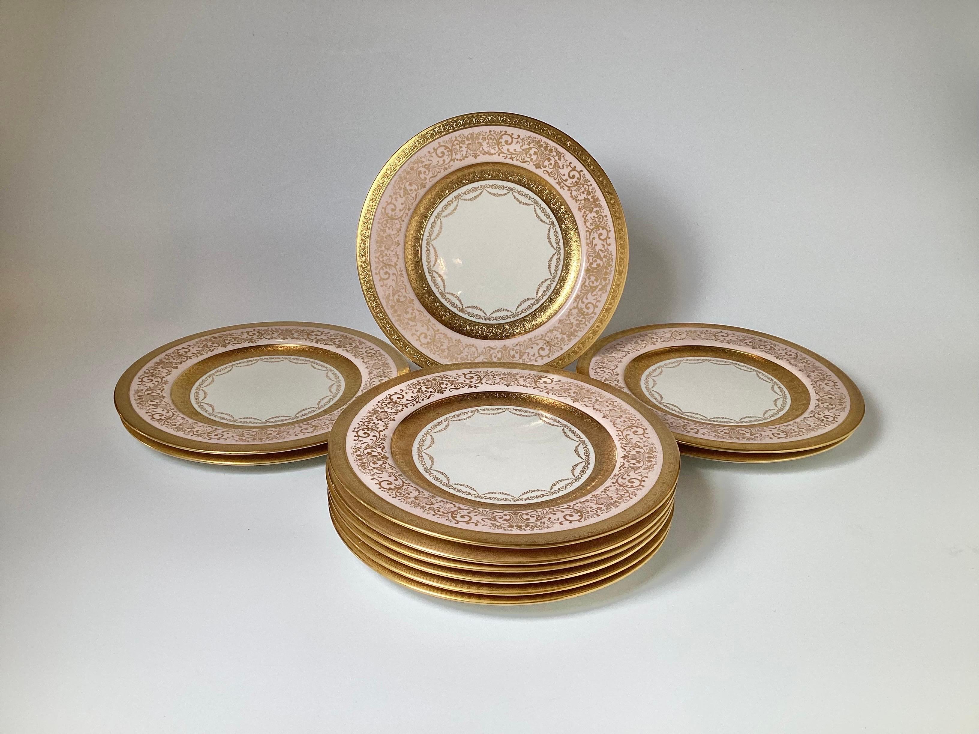 Elegant and formal gold decorated service plates with a pale pink border. The broad pink border with inner and outer gold band with gold scrolling detail with a swaged ring around the center white surface. These are beautiful plates with soft