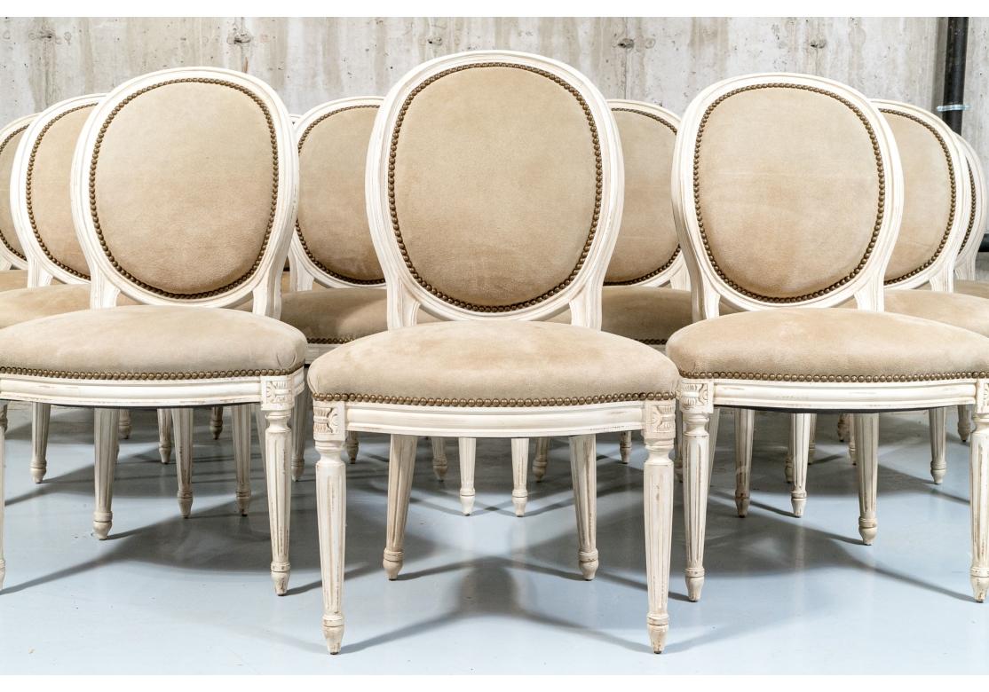 An elegant and well made set that are also quite comfortable. Set of 12 Louis XVI dining chairs with cream paint decorated frame and faux suede leather upholstery. The chairs with serpentine front, balloon back, carved wheat sheave medallions, brass