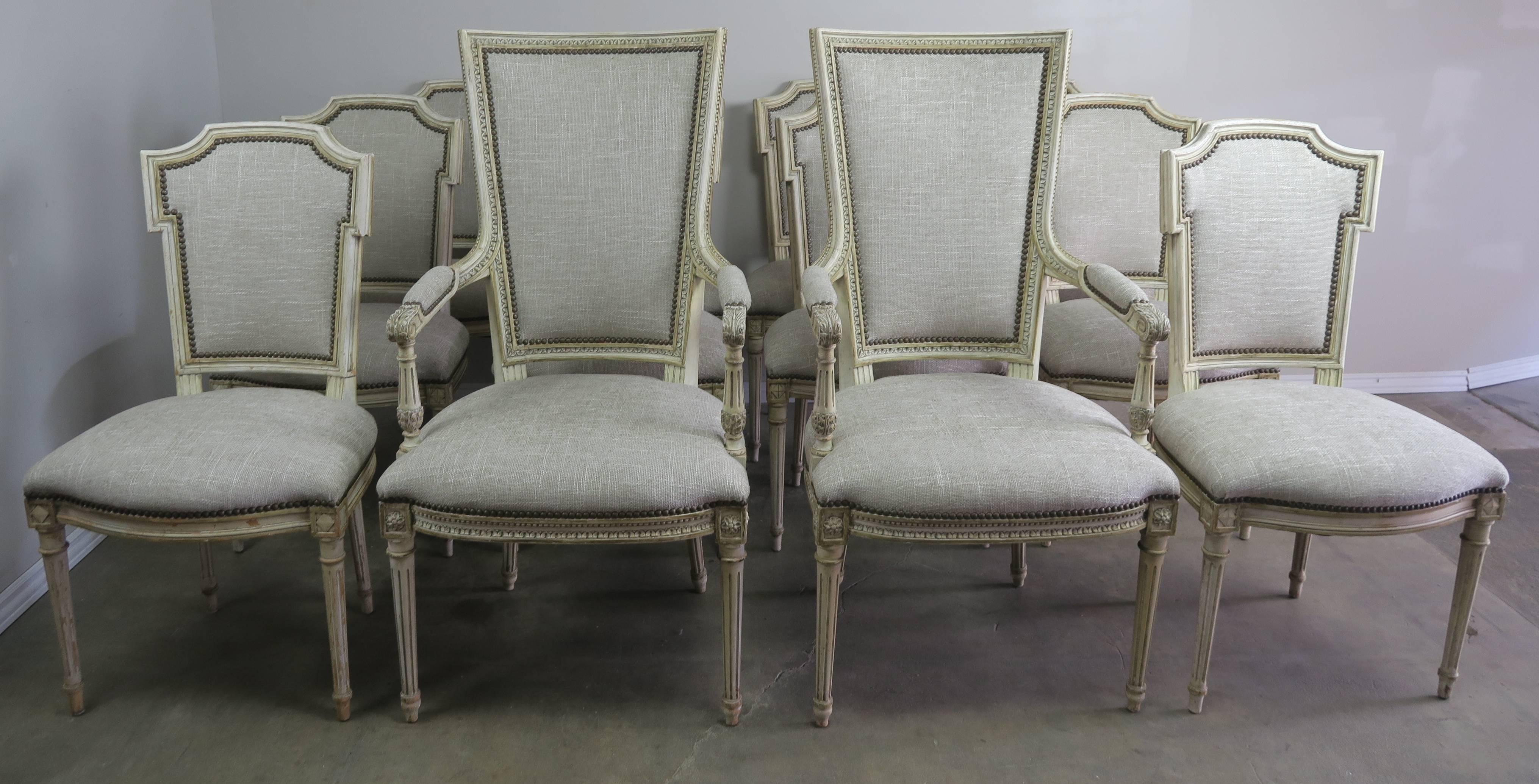 Set of 12 French Louis XVI style cream painted dining room chairs newly upholstered in linen with antique brass nailhead trim detail. The chairs each stand on four fluted straight legs. Size of large armchairs 24