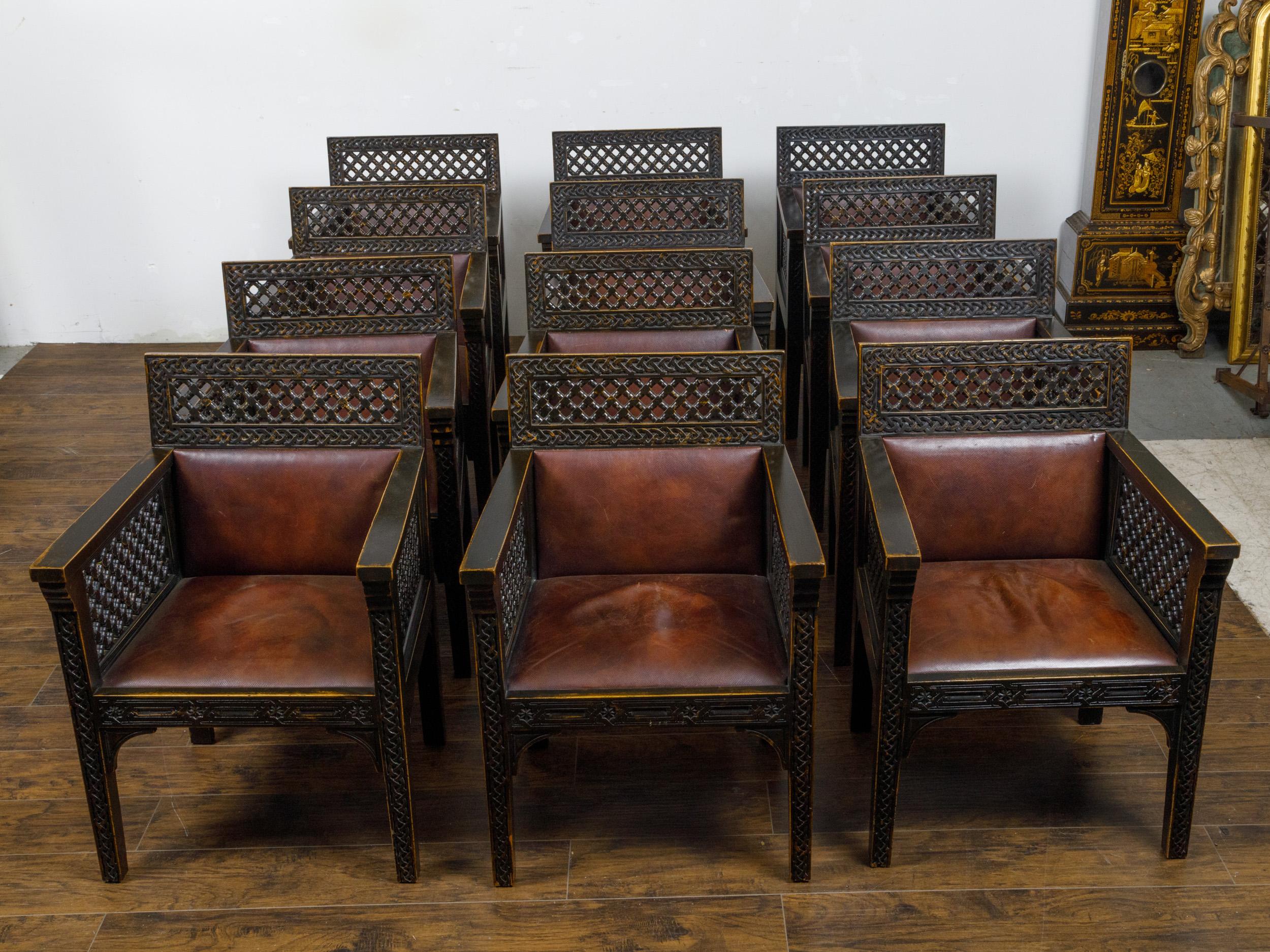 A set of 12 Moroccan ebonized wood armchairs from circa 1900 with carved décor and leather seats and backs. Crafted with an eye for detail, this set of twelve Moroccan ebonized wood armchairs from circa 1900 presents a harmonious blend of form and