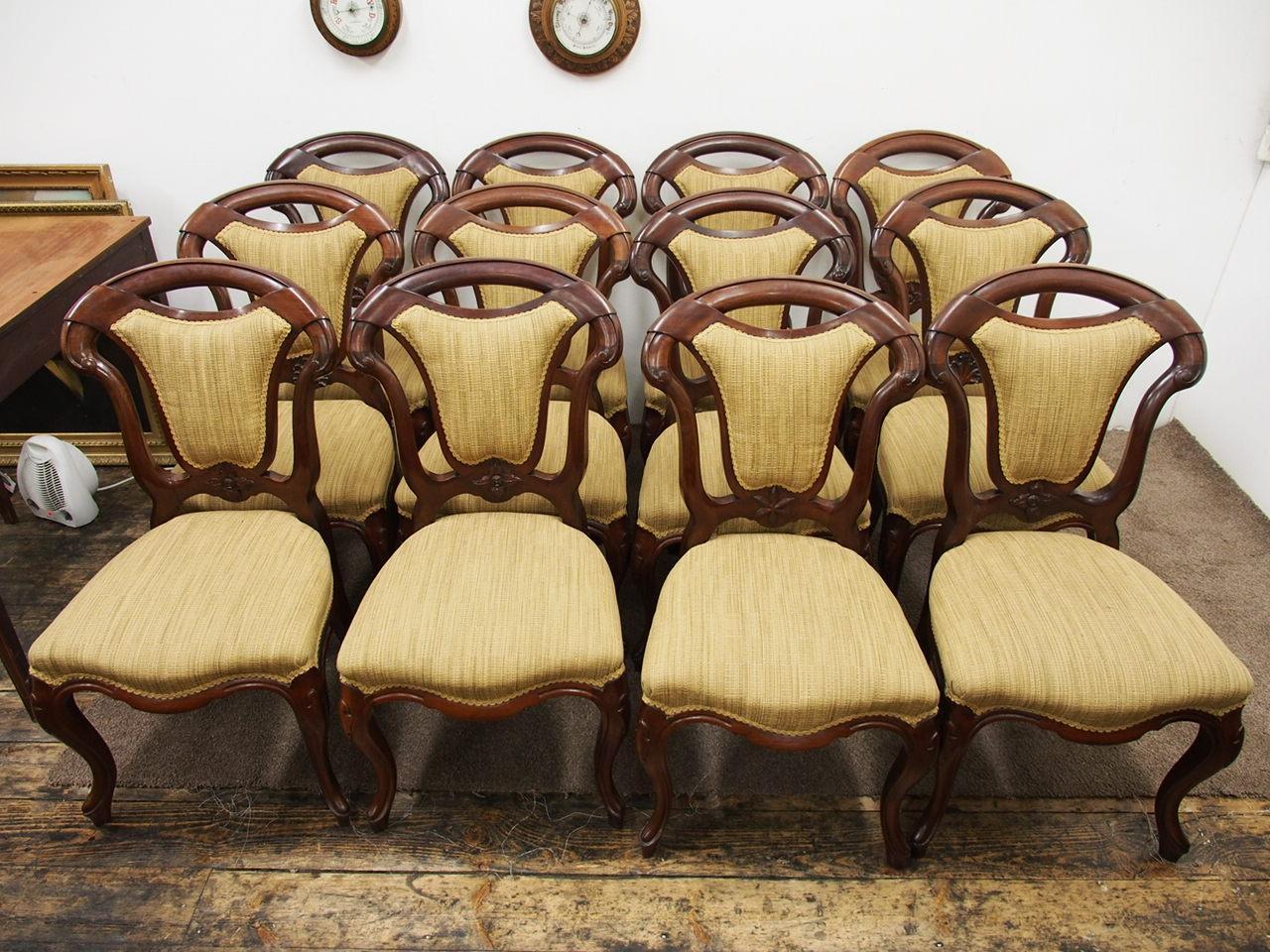 Set of 12 mahogany and upholstered Victorian dining chairs, circa 1870. The chairs have a curved top rail with a cartouche padded back and shaped uprights. At the base of the upholstered back is a carved leaf and the patterns vary from chair to
