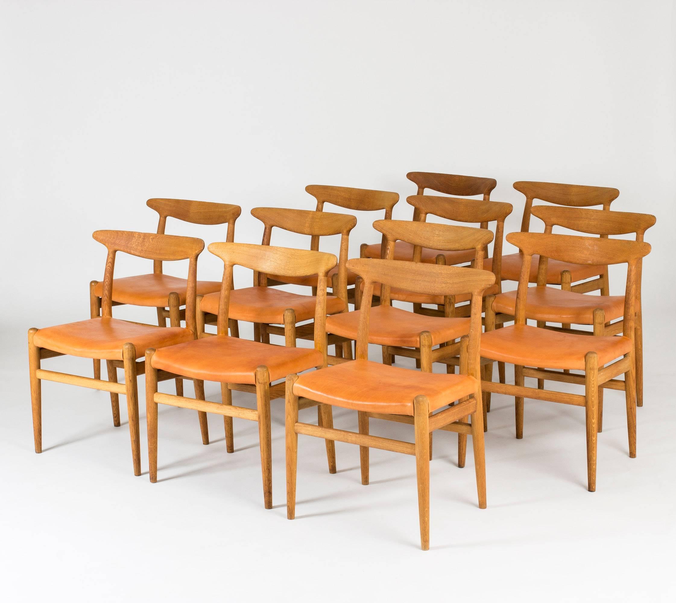 Set of 12 beautiful “W2” dining chairs by Hans J. Wegner, made from oak with cognac leather seats. Great woodgrain that follows the lines of the seamlessly sculpted backrests.