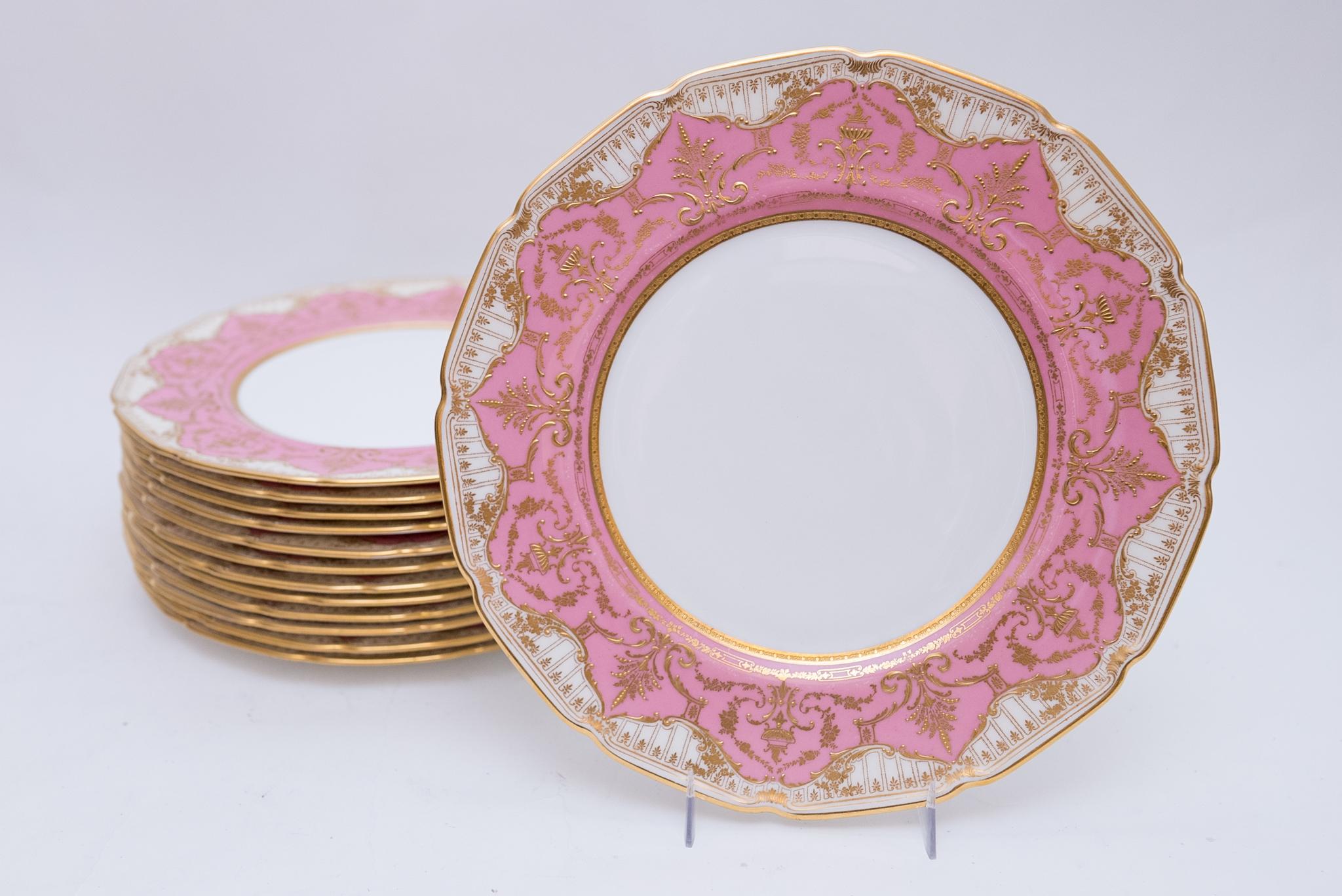 A pretty set of pink and raised gilt dinner plates, a set of 13 with one extra. This set features a collar of pink and light cream swirl pattern with finely detailed gilding in an urn and floral design. In wonderful antique condition and ready to