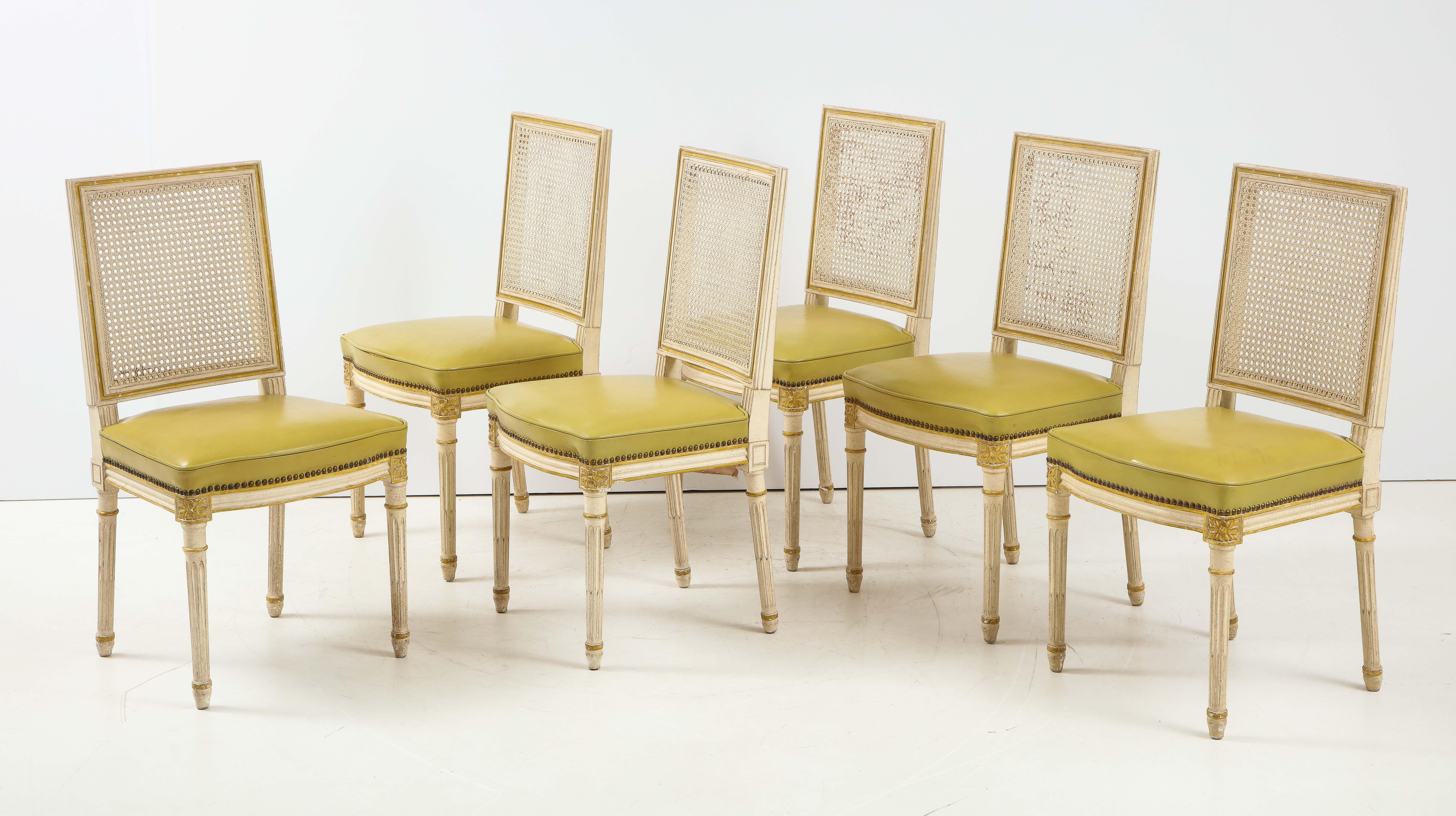 A set of stylish dining chairs made by Maison Jansen, Paris. The Lous XVI style chairs are painted in a washed white finish with subtle accents in a chartreuse color. The chairs feature a caned back and seats finished in a green faux leather trimmed